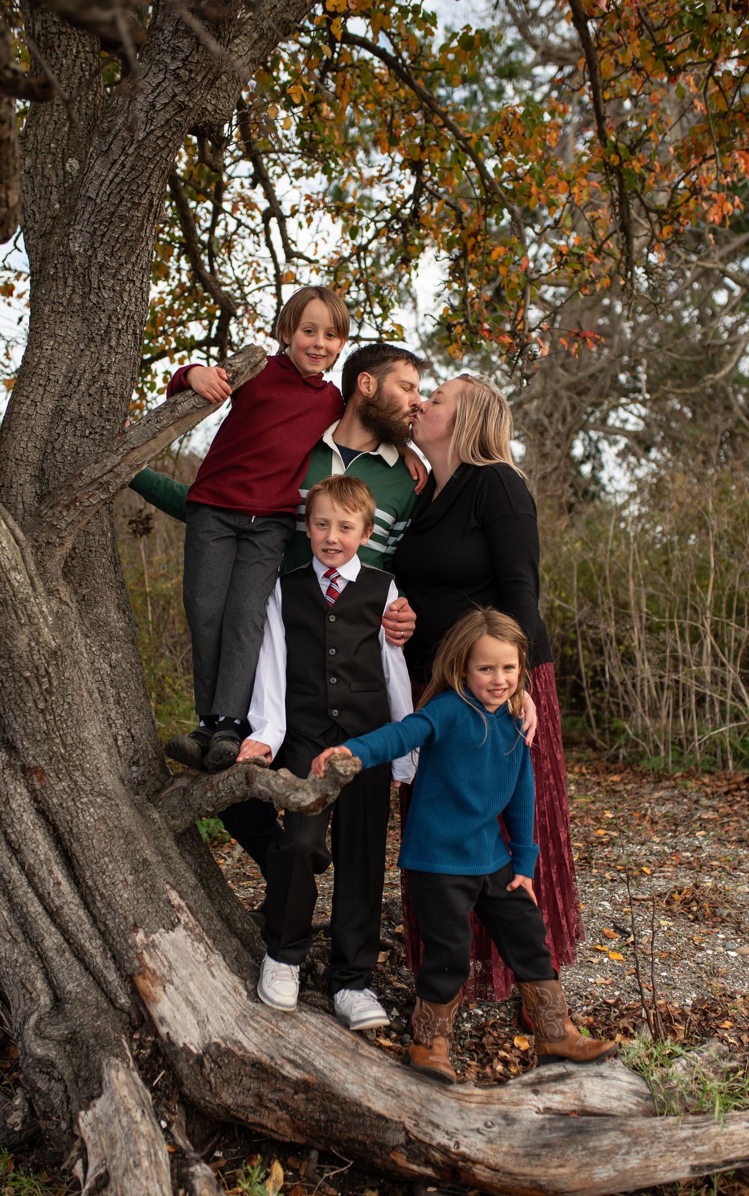 Family of five posing on large tree roots in autumn forest, parents kissing.
