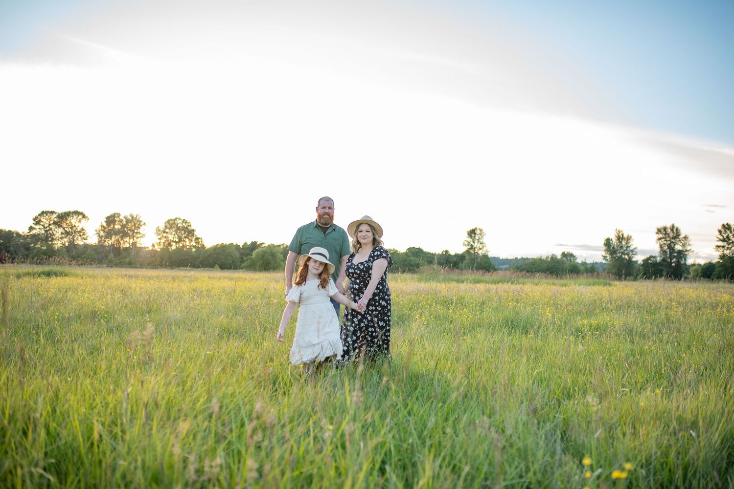 Family of three stands together in a wide green meadow during a beautiful sunset.
