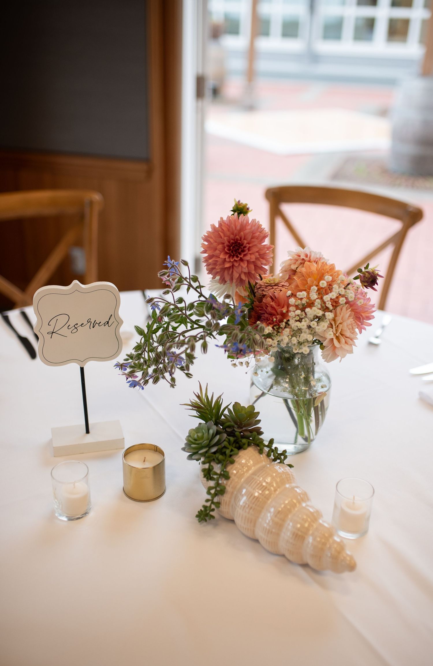Elegant wedding table centerpiece with pink dahlias, succulents, seashell, candles, and a reserved sign on white linen.
