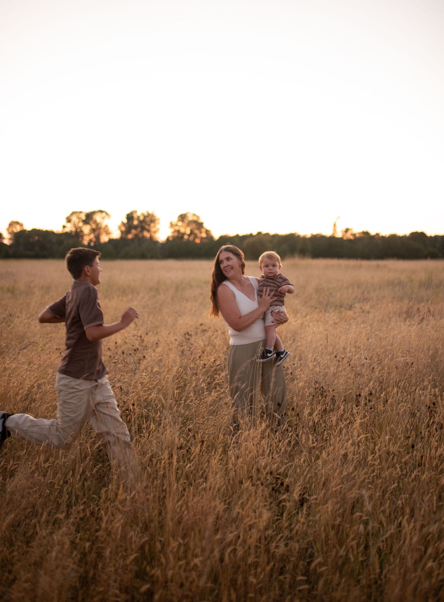 Family walking through golden wheat field at sunset, mother holding baby while child runs nearby.