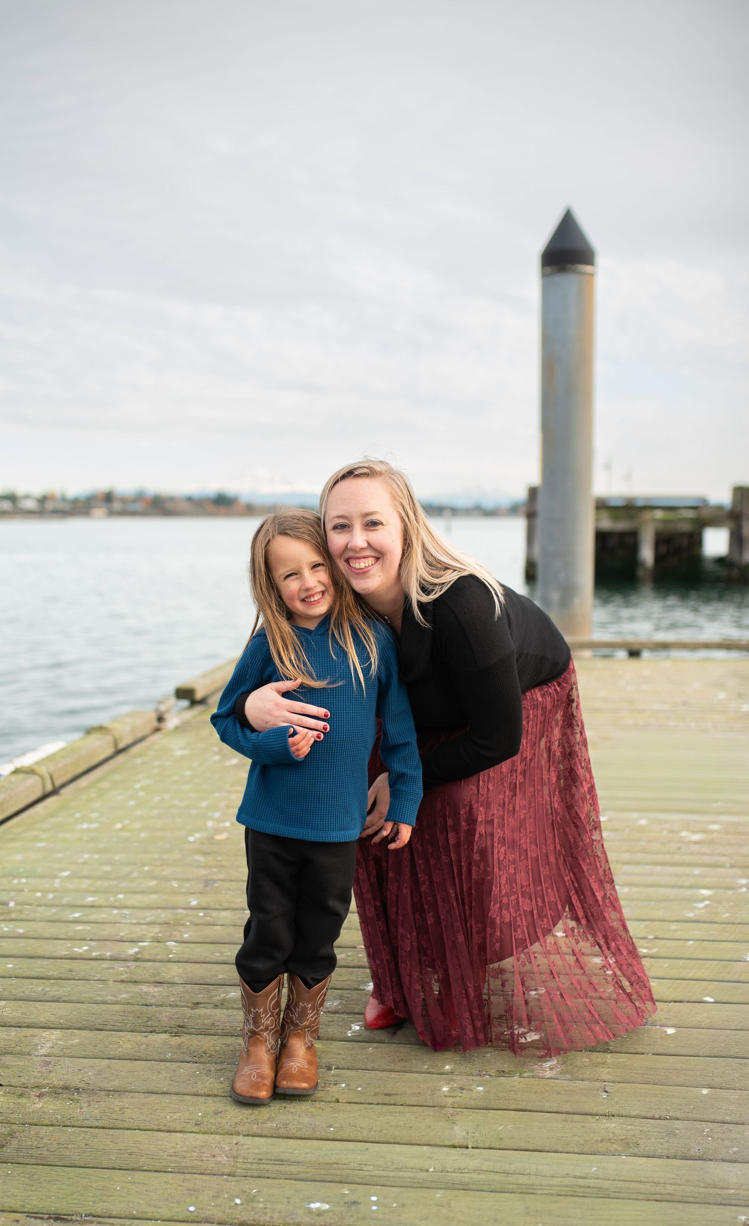 Blonde mother hugging young child in blue sweater on wooden dock by water.