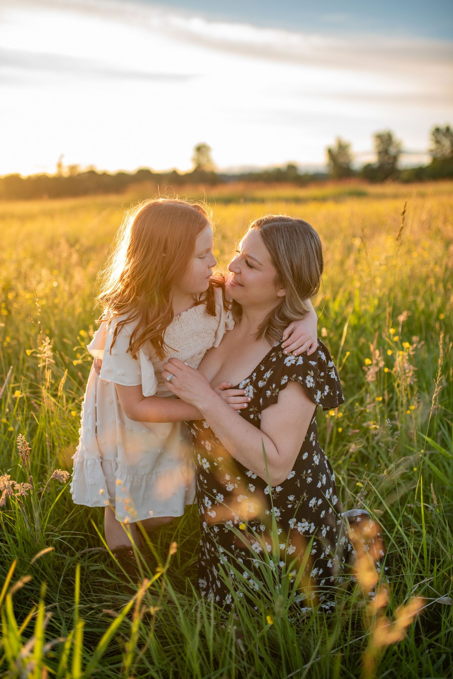 Mother and red-haired daughter share a loving face-to-face moment in a golden sunlit field.