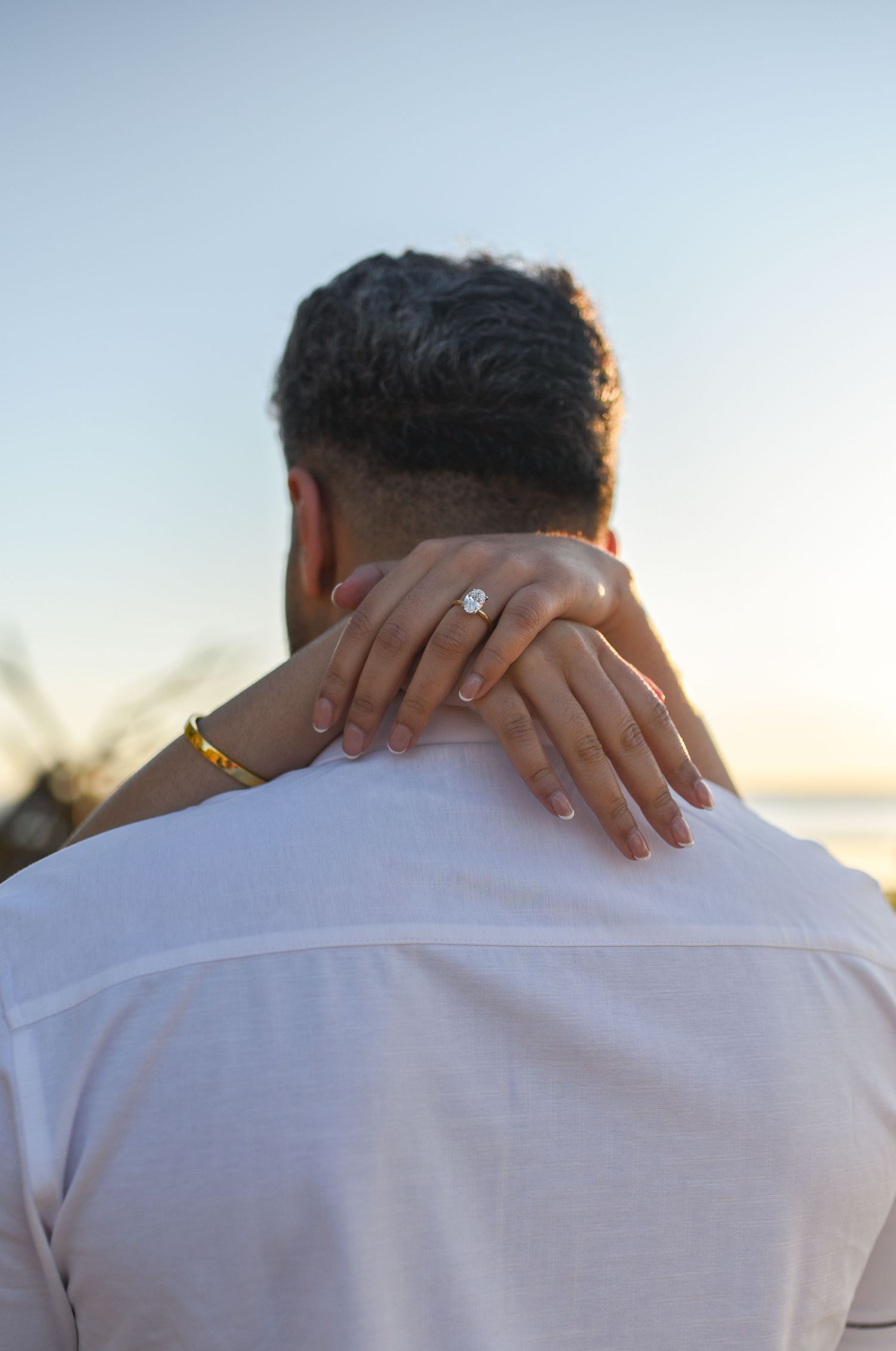 Close-up of engaged woman's hand with diamond ring resting on man's shoulder at sunset beach.