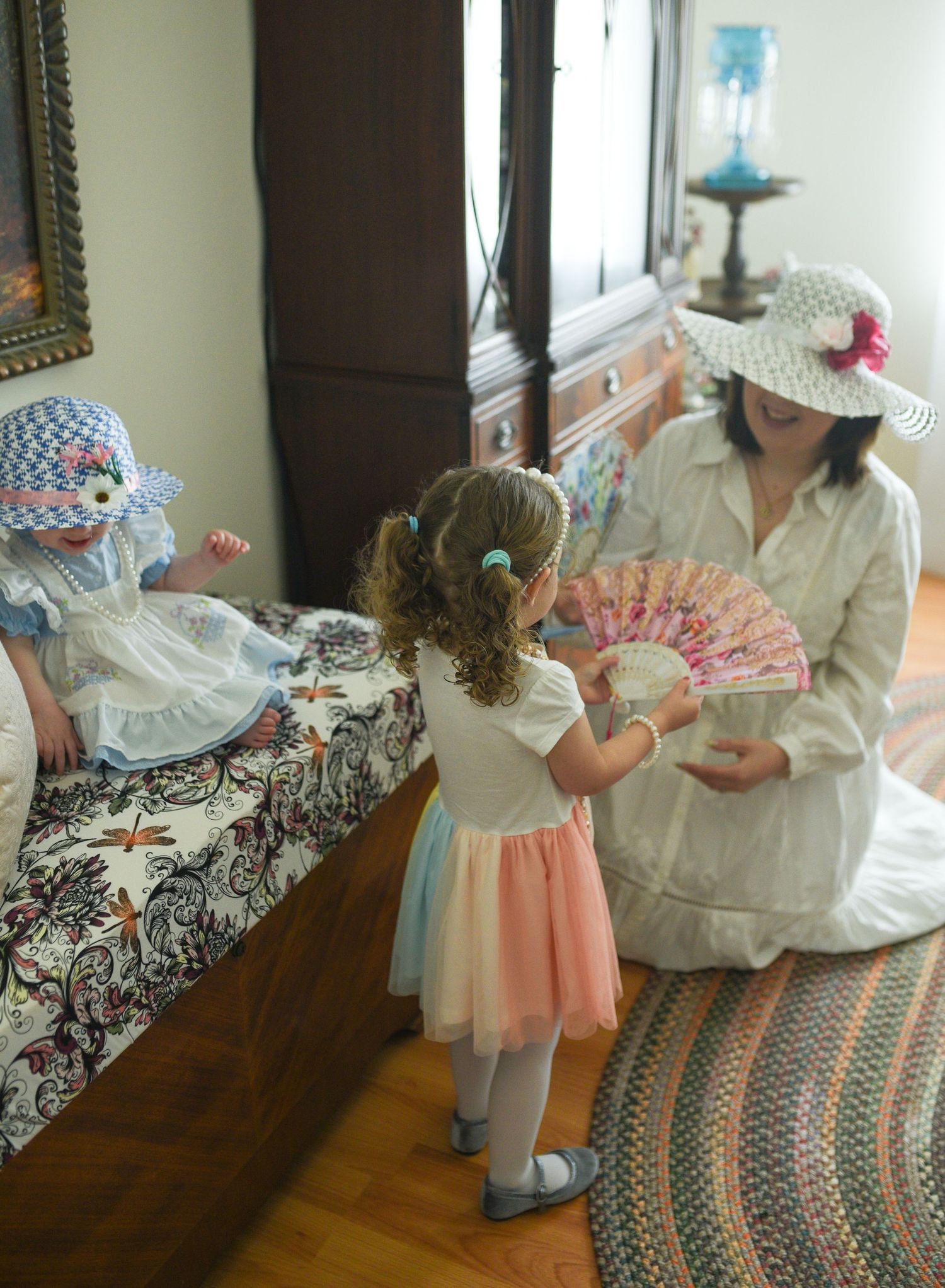 A woman and two young girls in vintage hats enjoy a tea party in a charming bedroom setting.
