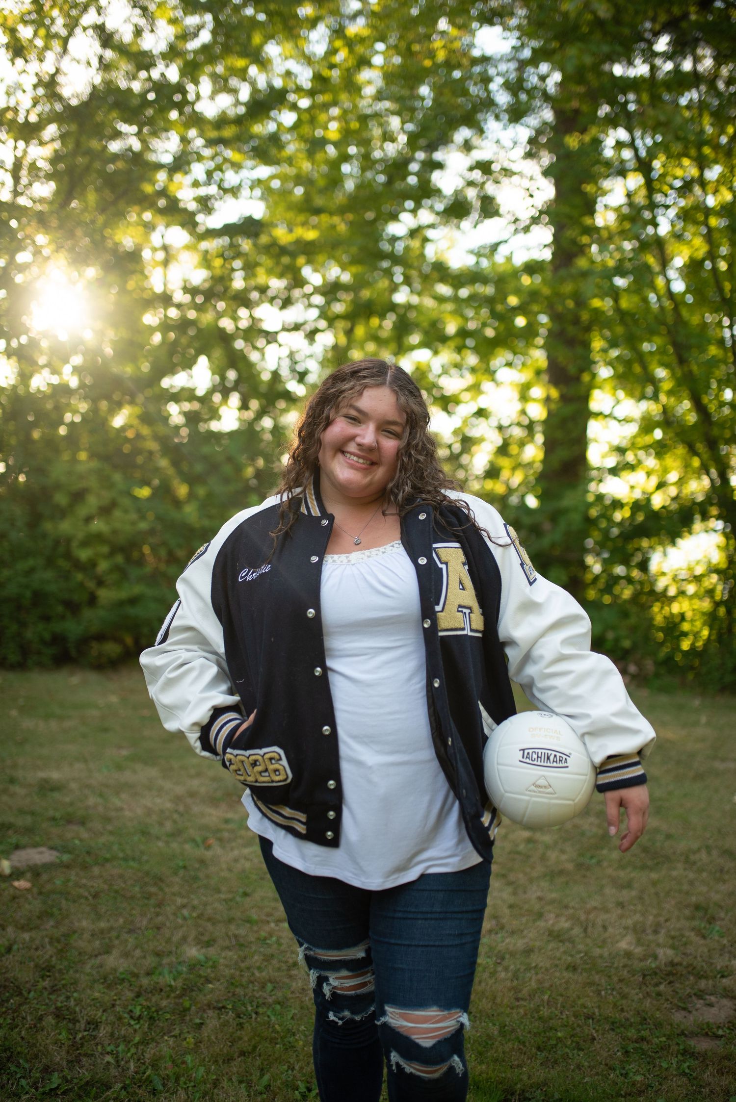 Smiling teen girl holds volleyball wearing varsity letterman jacket outdoors.