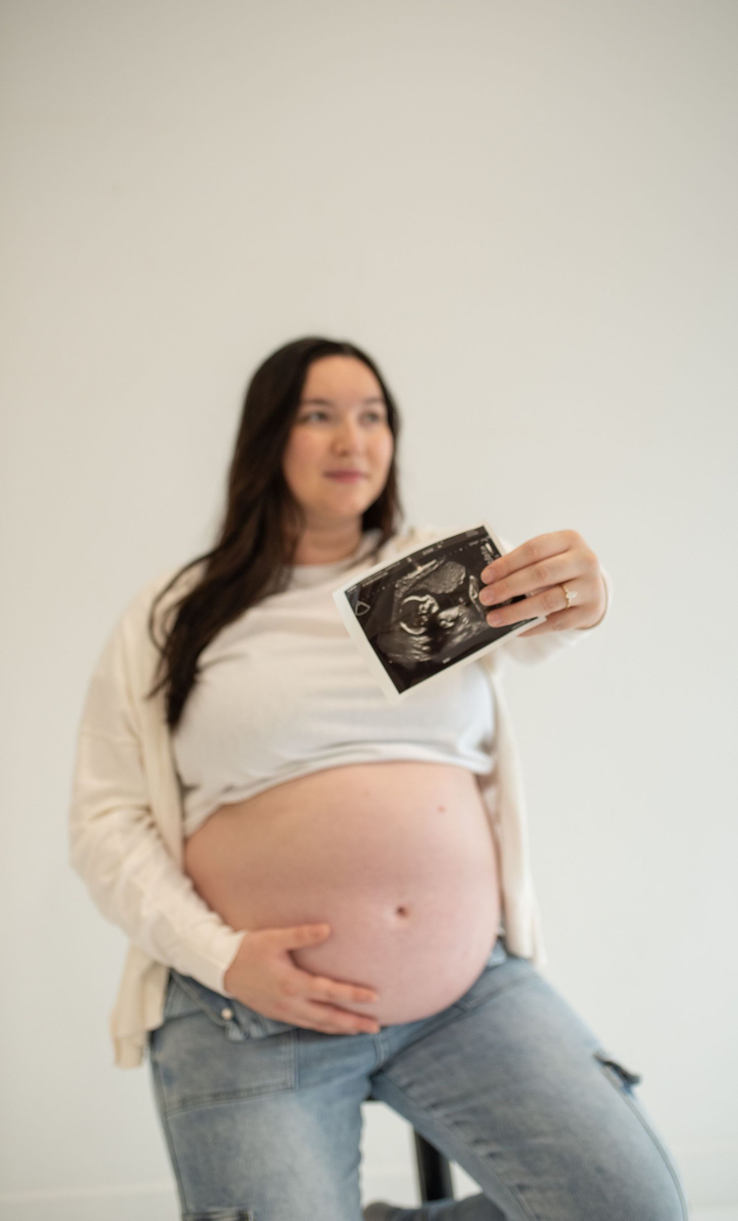 Pregnant woman in white cardigan holding ultrasound photo while cradling her bare baby bump in a bright white studio.