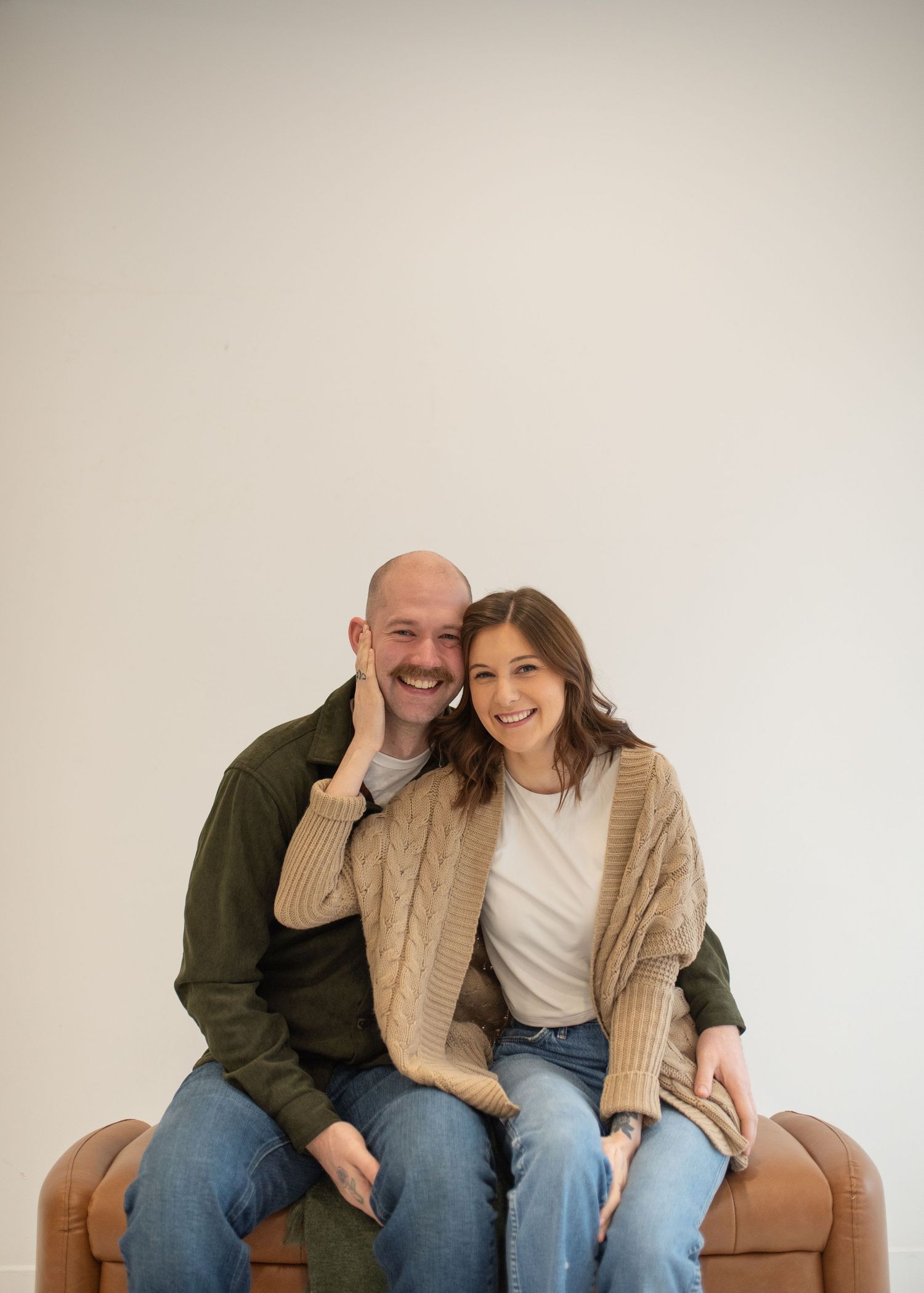 Smiling couple sitting together on leather bench against white wall in cozy indoor portrait session.