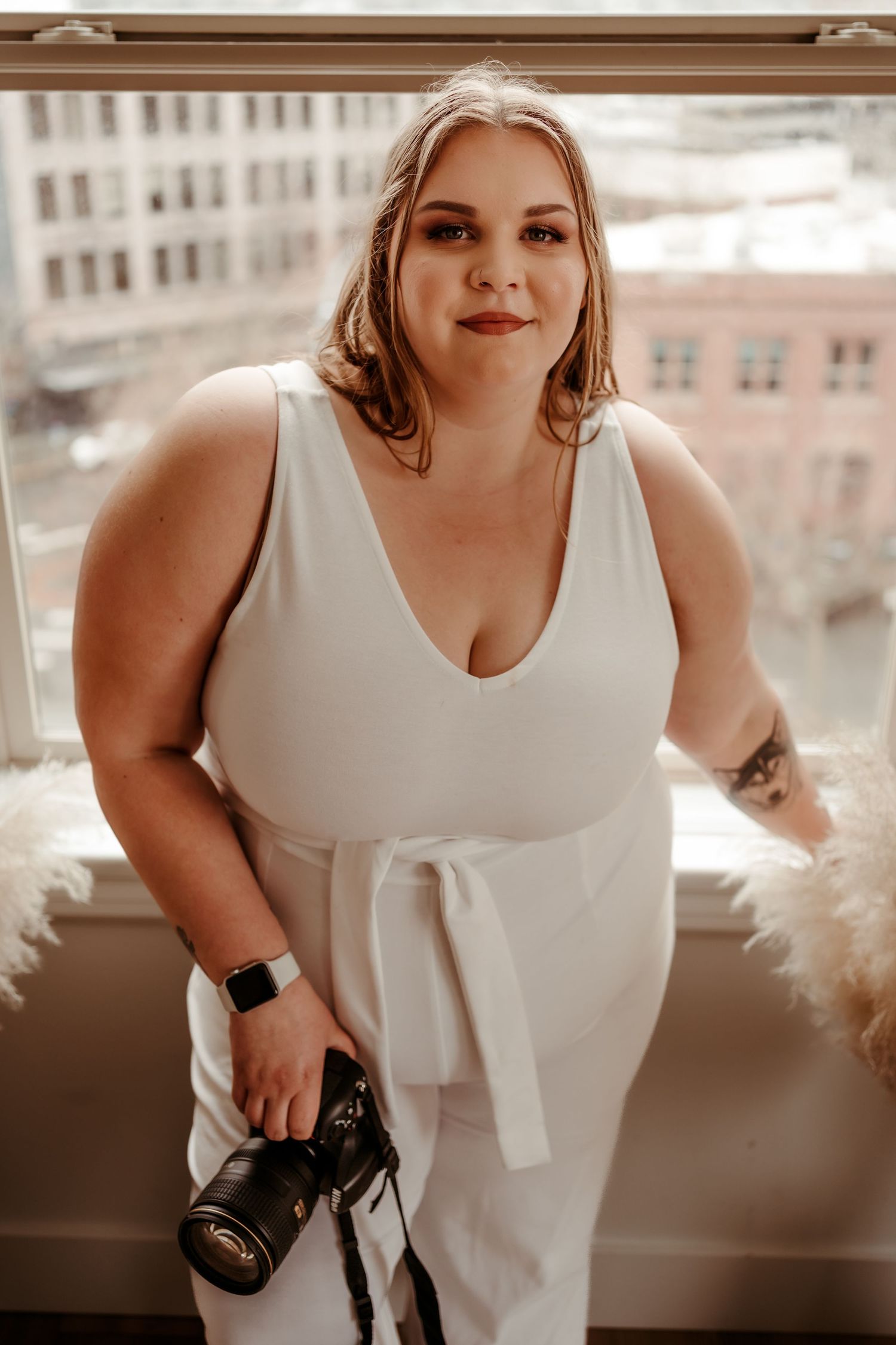Woman in white jumpsuit holding a DSLR camera while standing by a window.