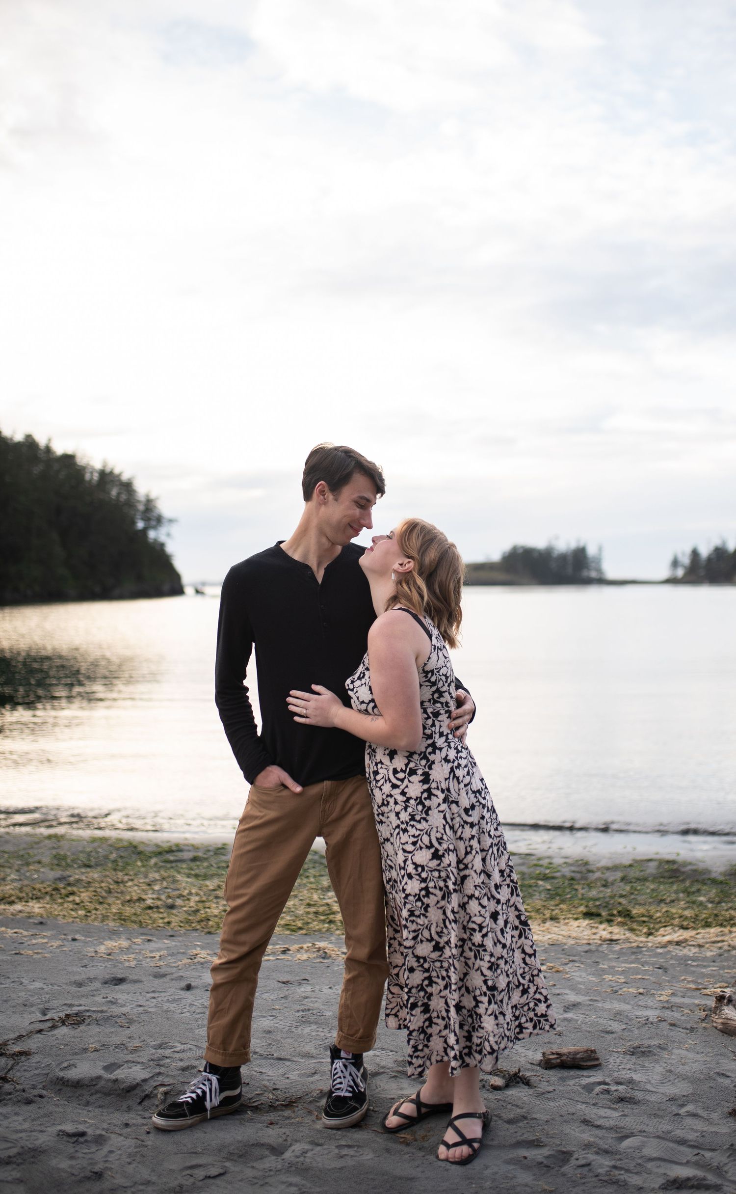 Couple sharing a romantic moment on a rocky beach, man in black top and woman in floral dress by calm water.