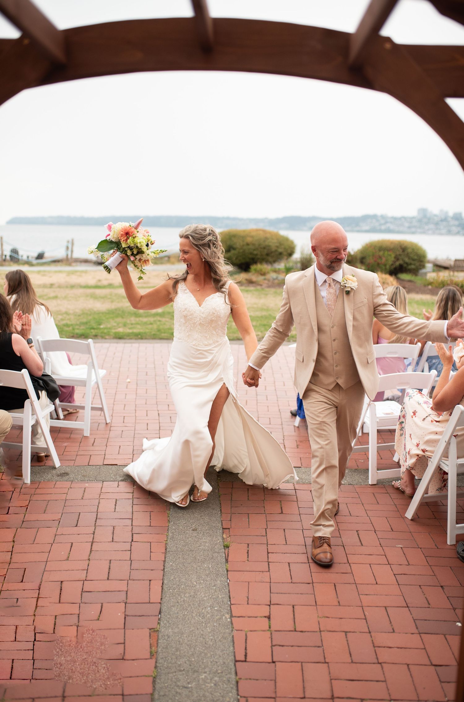 Bride and groom walk down brick aisle after outdoor wedding ceremony, bride holding colorful bouquet aloft.