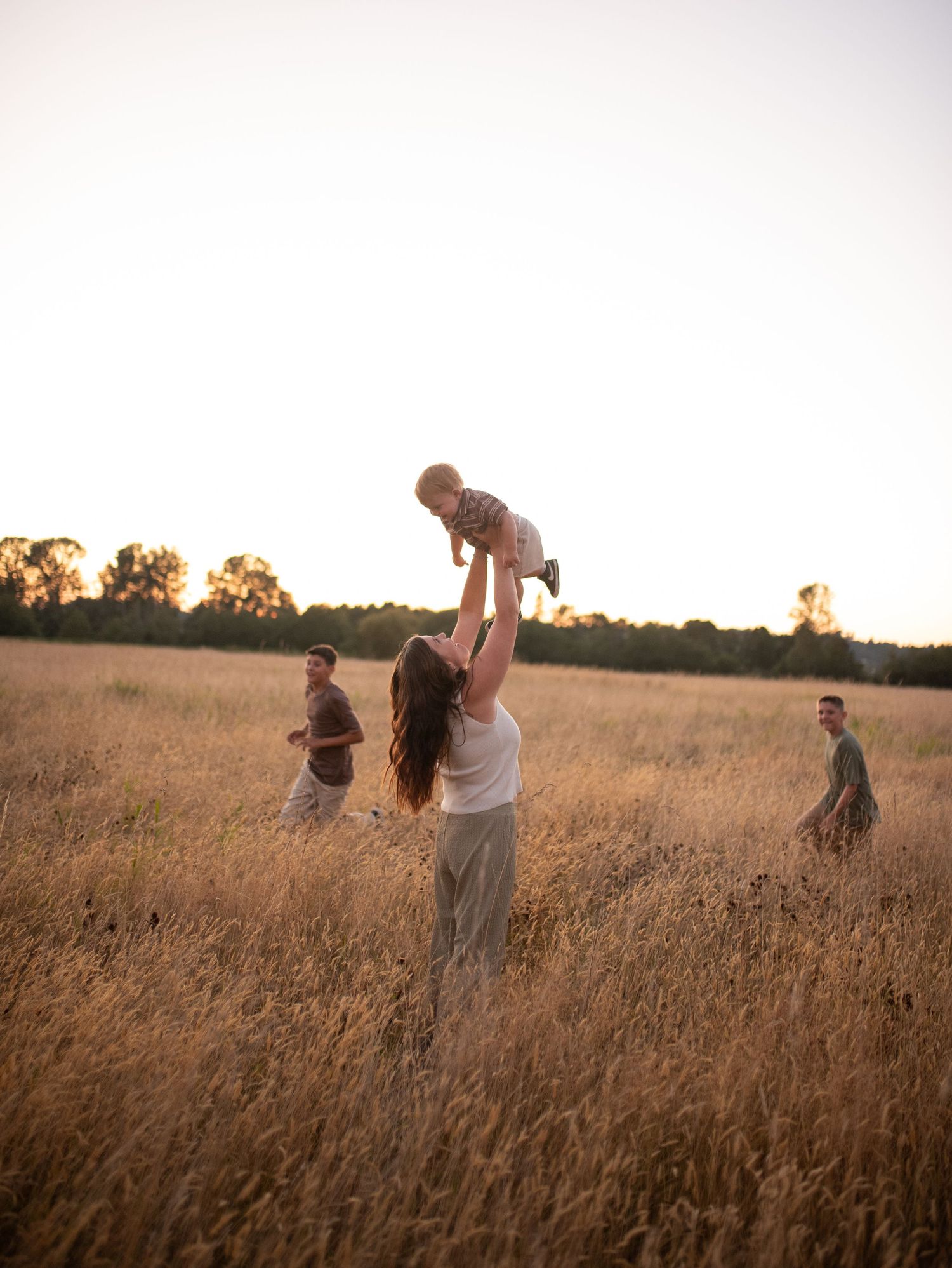 Mother joyfully lifting baby overhead in golden field at sunset with family nearby.
