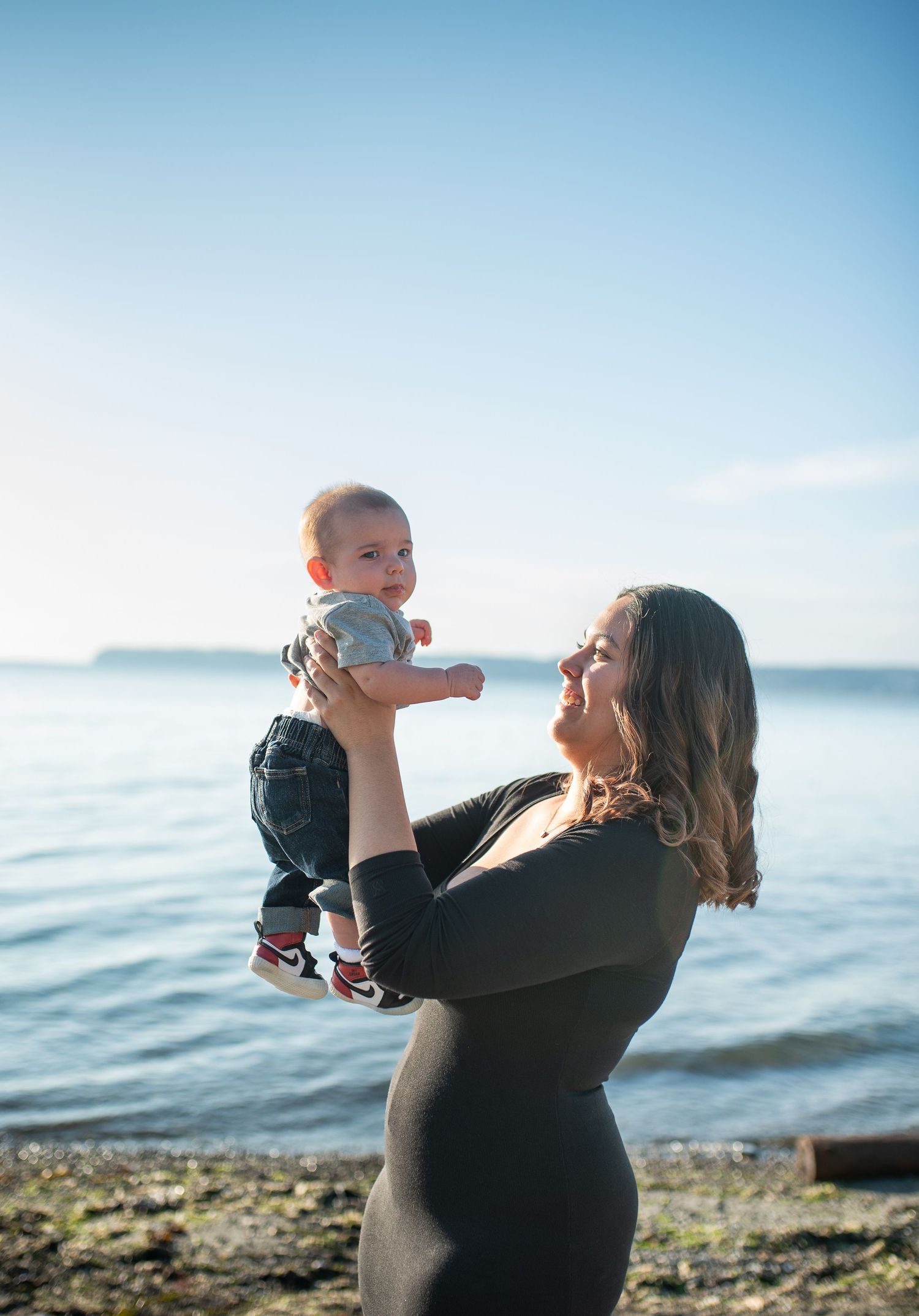 A mother joyfully lifts her baby by the ocean shore on a sunny day, both smiling in a heartwarming moment.