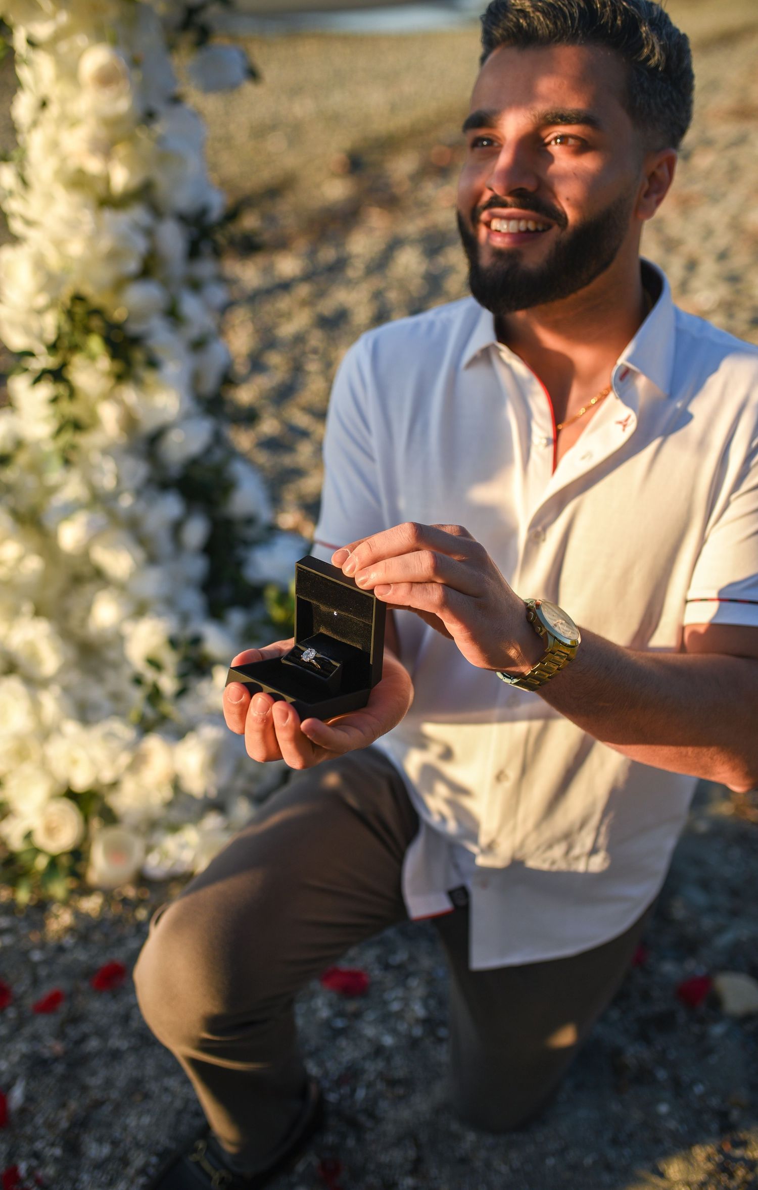 Man kneeling on beach holding open engagement ring box surrounded by white roses during golden hour proposal.