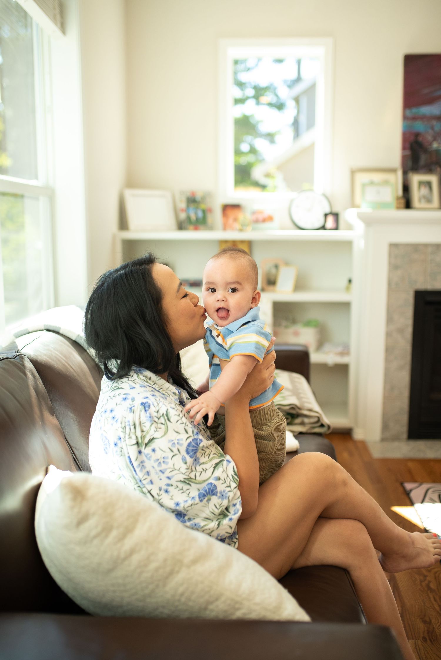 A mother kisses her laughing baby while sitting on a leather sofa in a bright, cozy living room.