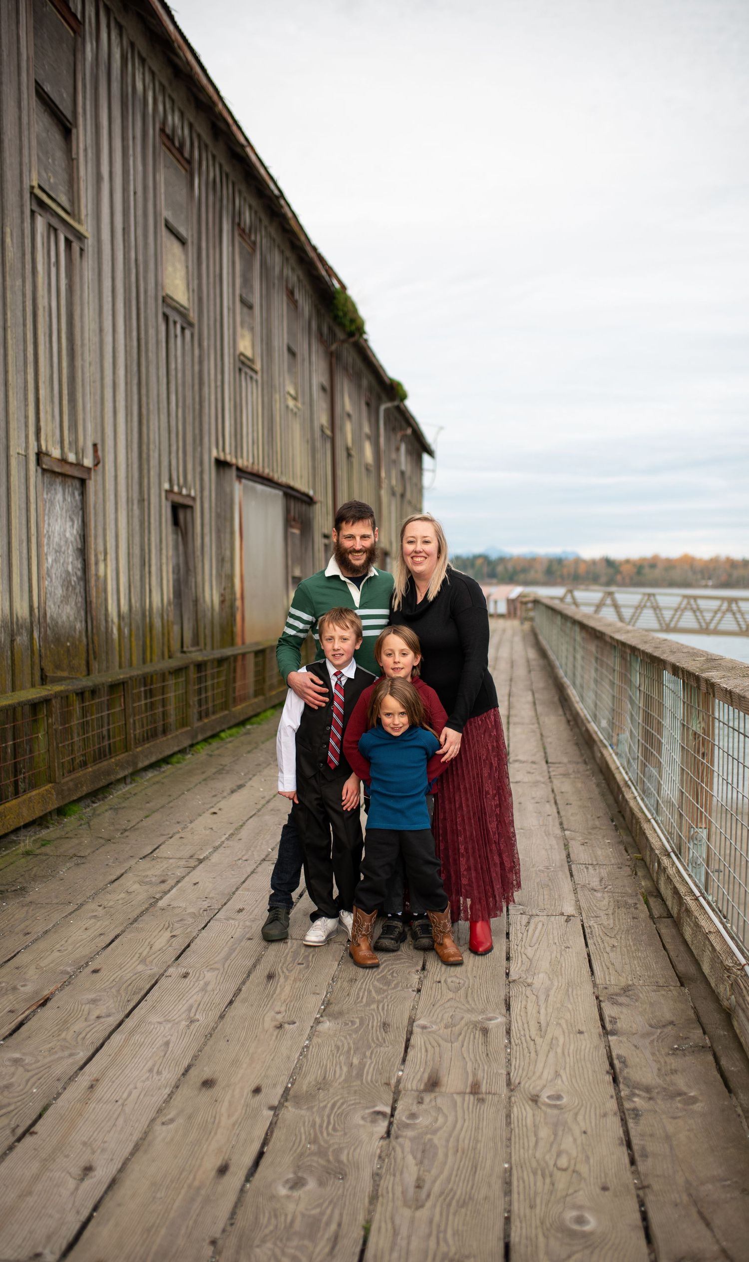 Family of five posing together on wooden boardwalk beside rustic waterfront building.