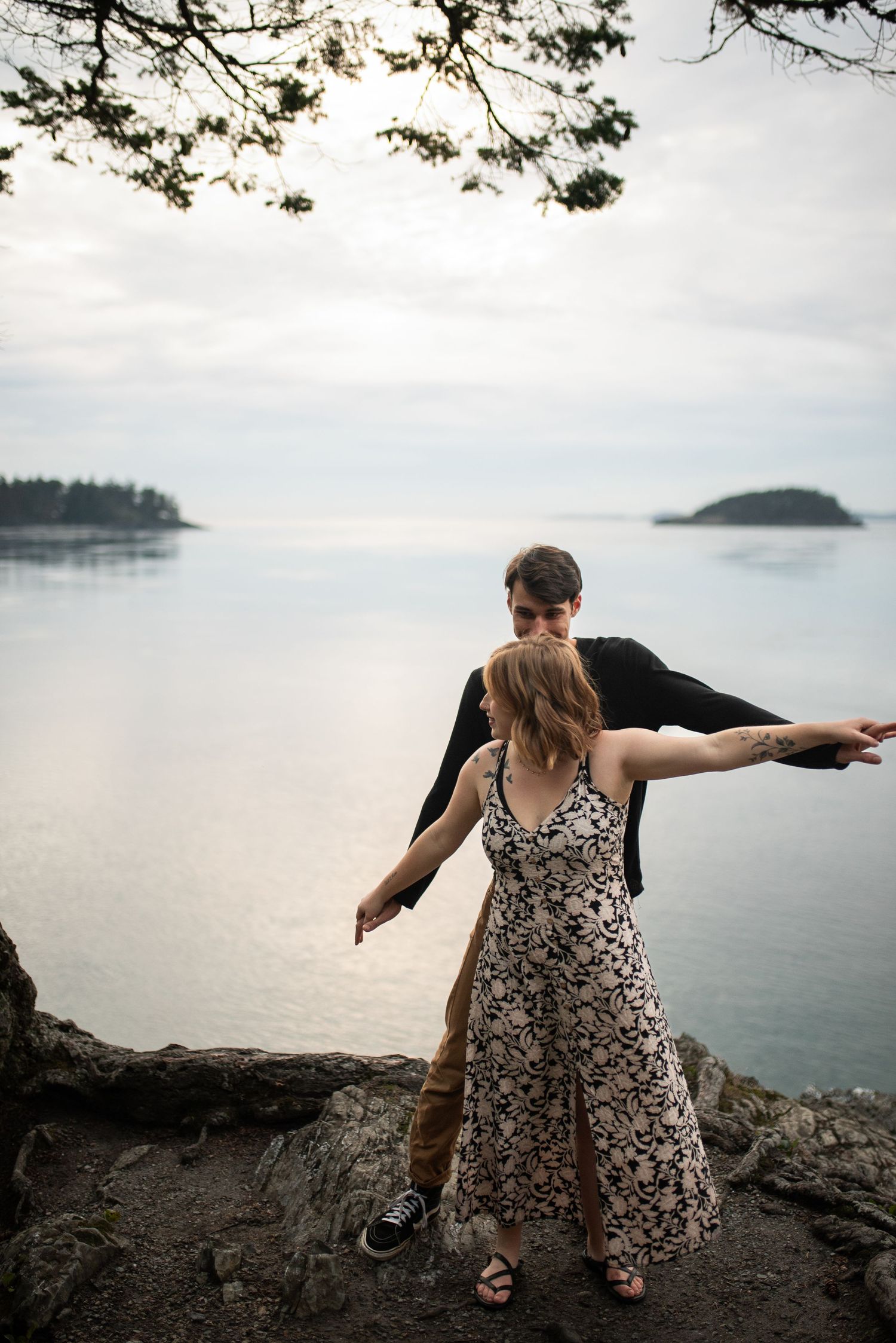 Couple balancing on coastal rocks beneath pine branches, arms outstretched over a serene ocean vista.