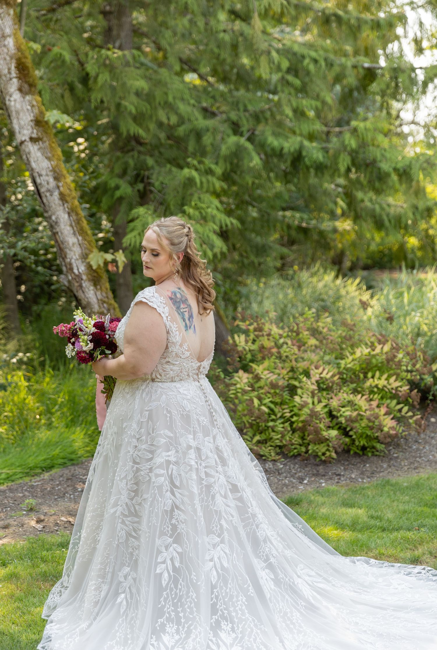 Bride in lace A-line wedding gown with floral tattoo, holding burgundy bouquet in lush garden setting.