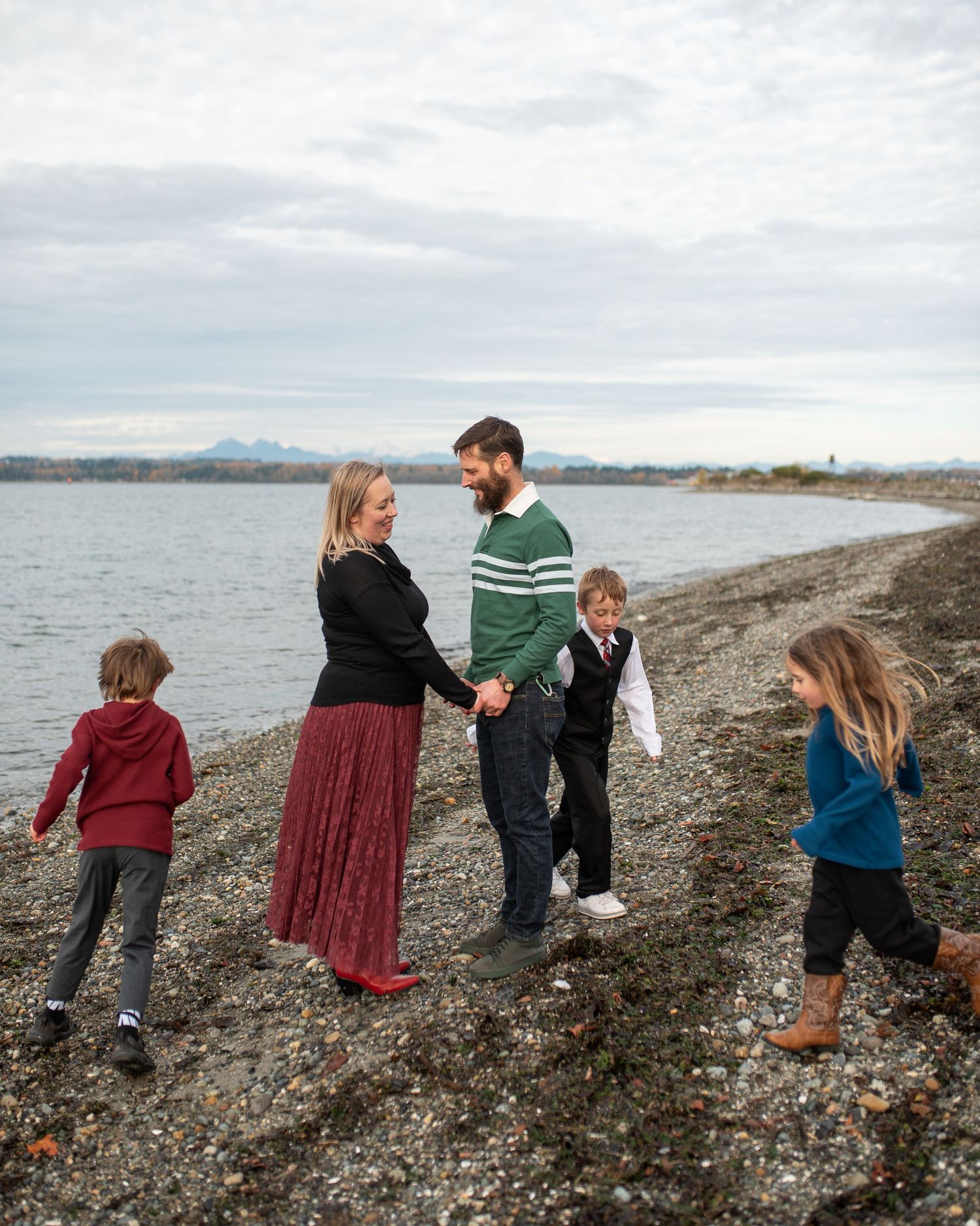 Family of five walking and holding hands on rocky beach shoreline on overcast day.