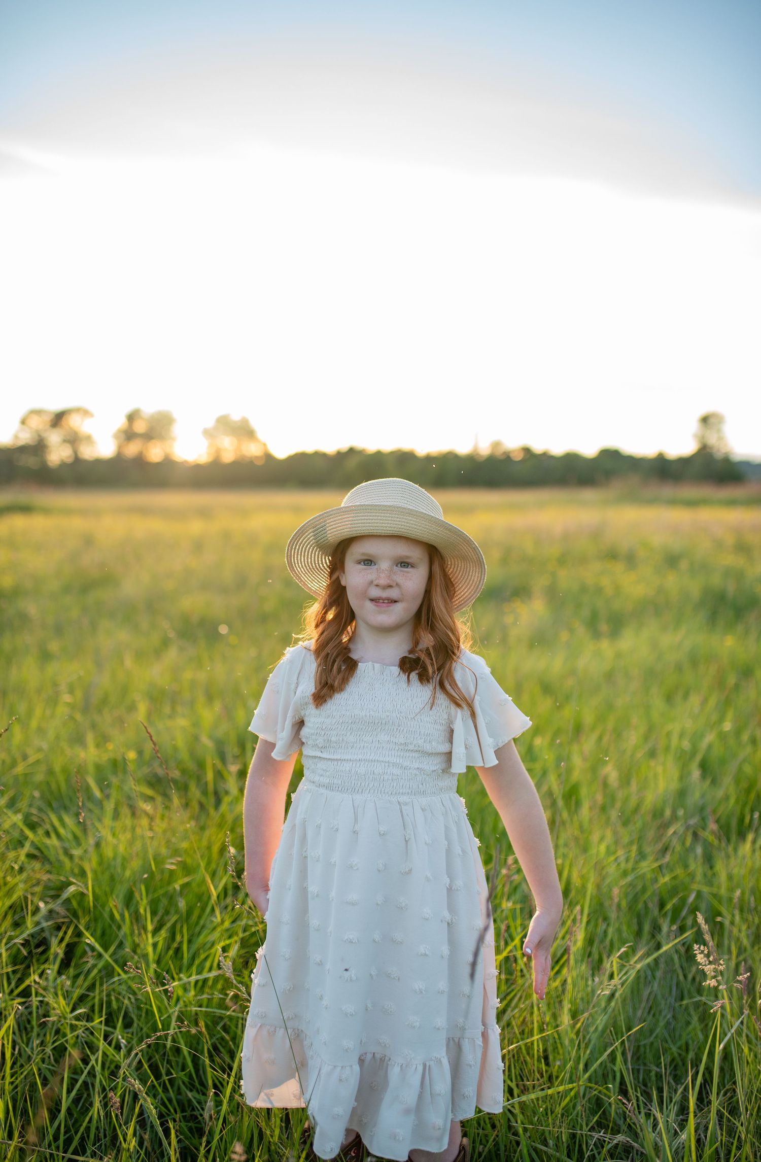 Smiling red-haired girl in white dress and straw hat stands in a sunlit green field.