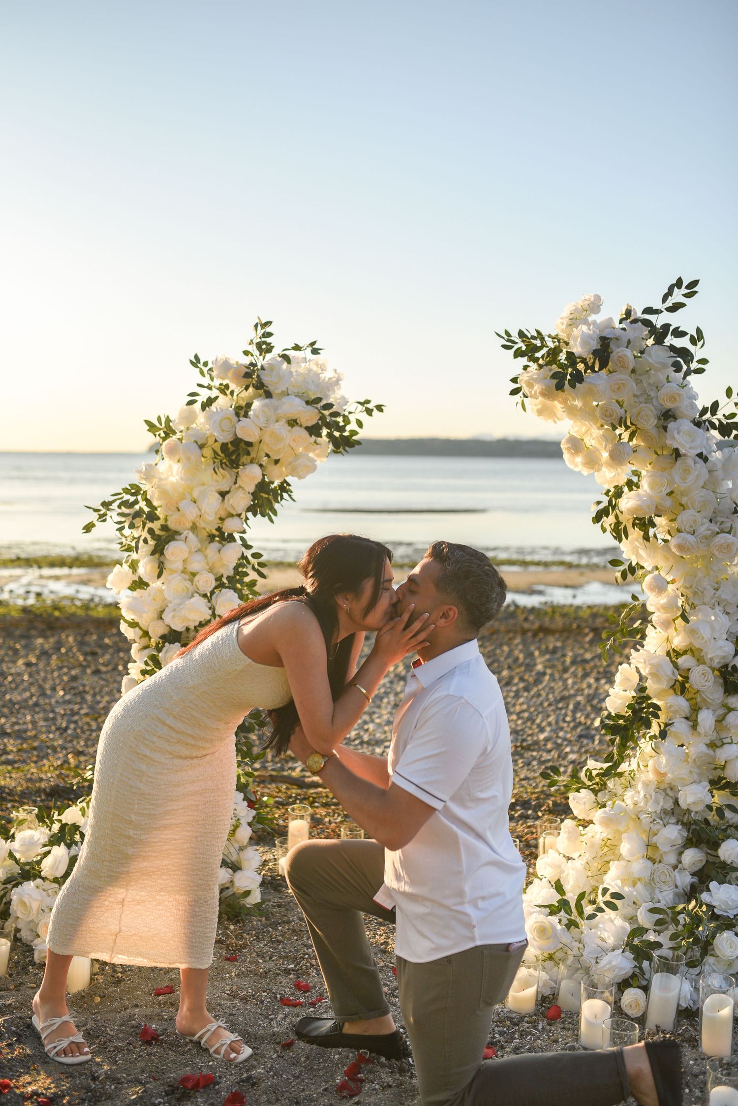 Newly engaged couple sharing their first kiss on a beach surrounded by white floral arches and candles.