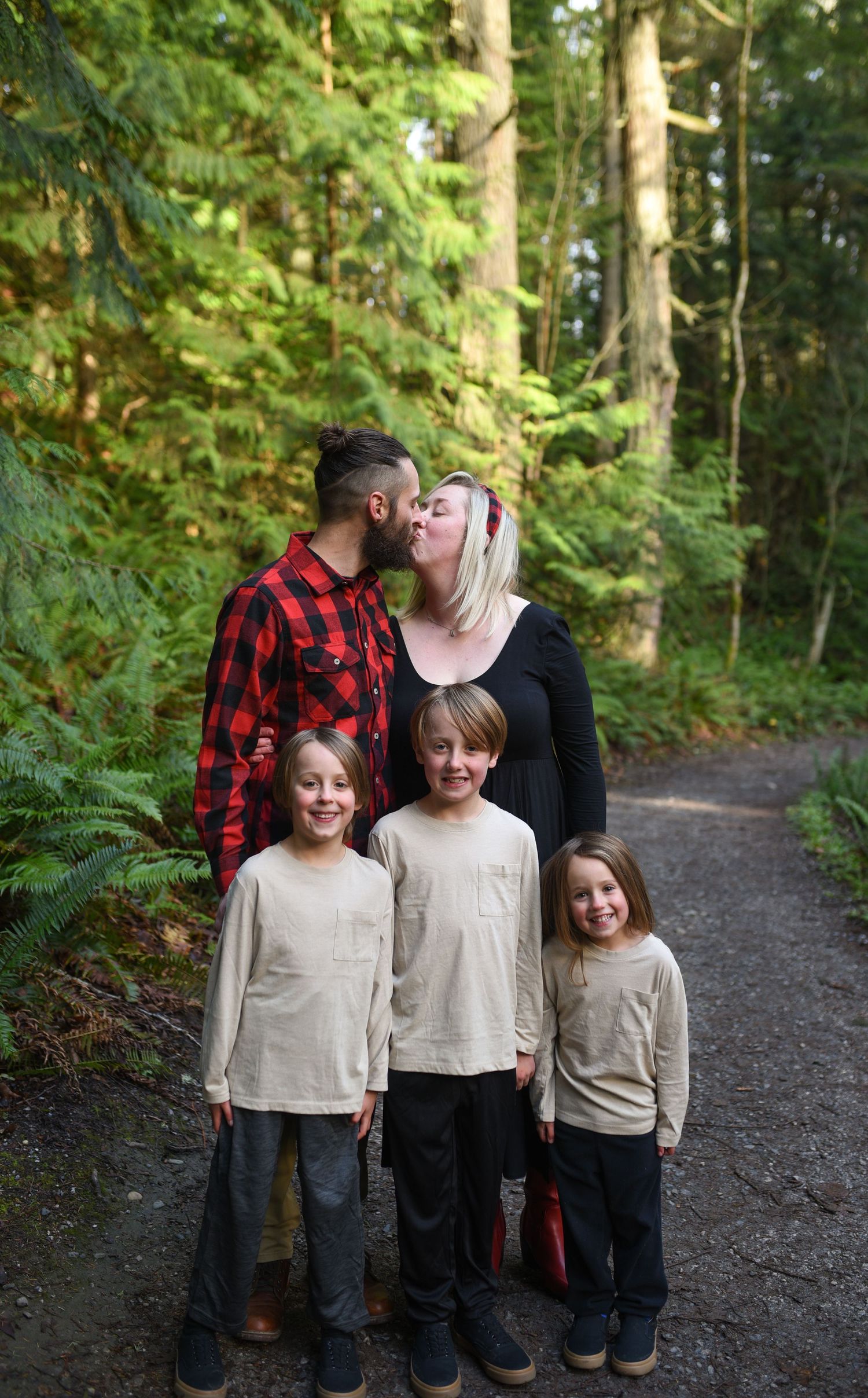 Family of five poses on forest path, parents kissing while three children stand in front.
