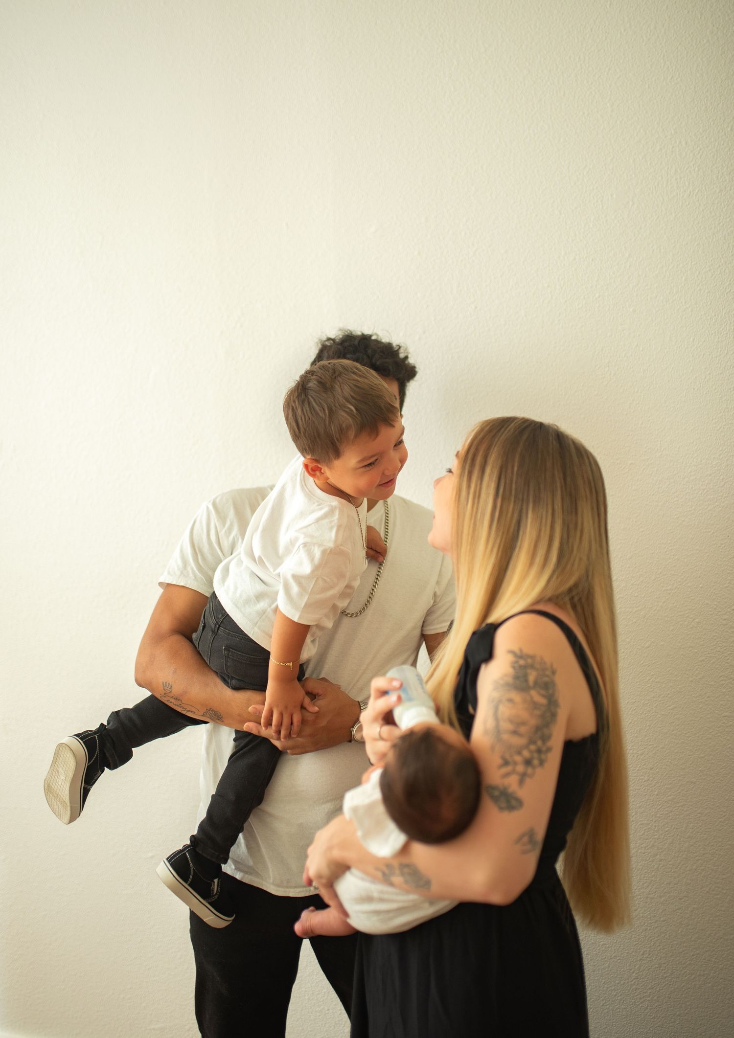 Father lifts toddler son while mother holds newborn baby in bright minimalist indoor setting.