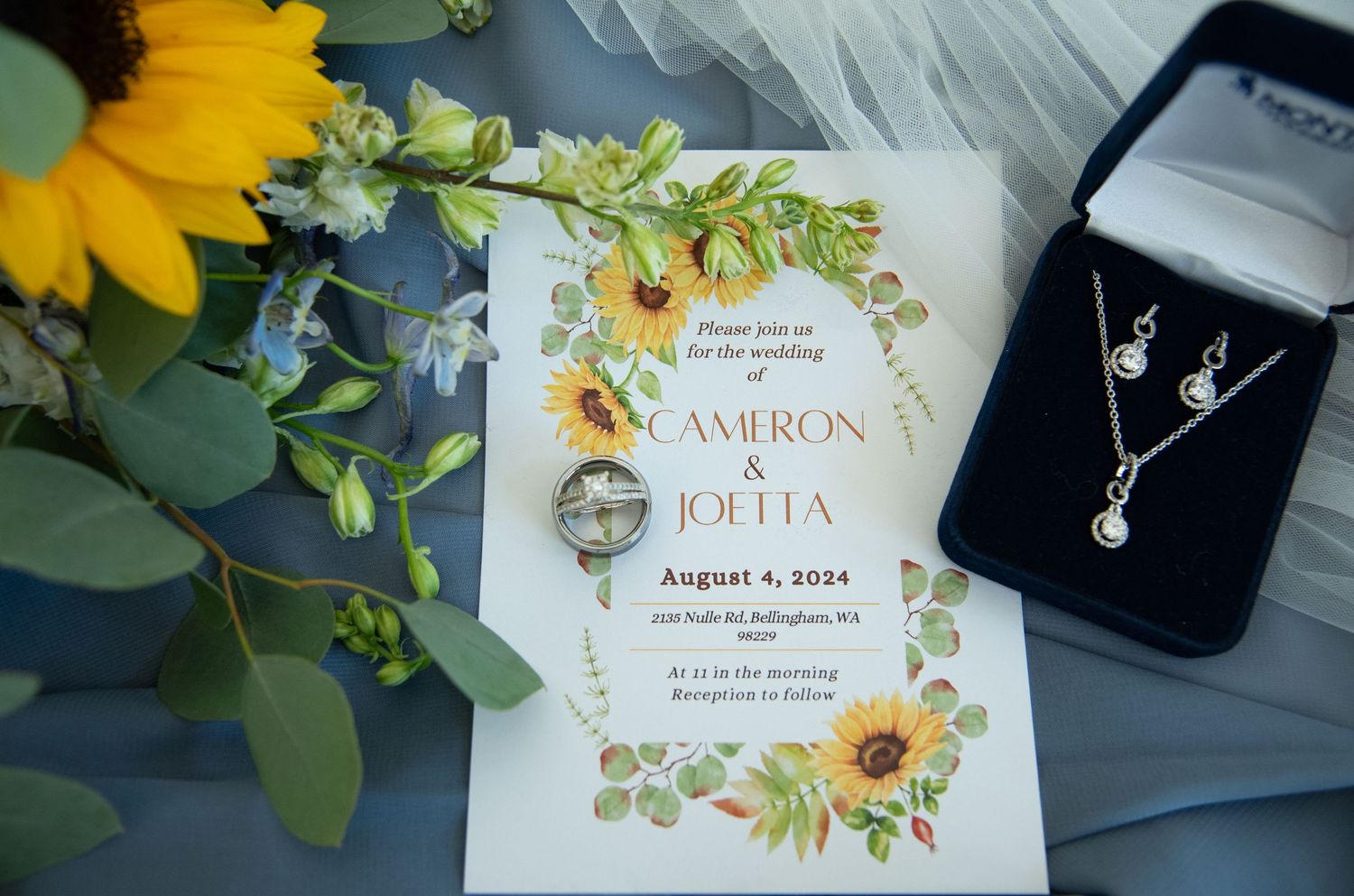 Wedding invitation for Cameron and Joetta on August 4, 2024, displayed with sunflowers, jewelry, and wedding rings.
