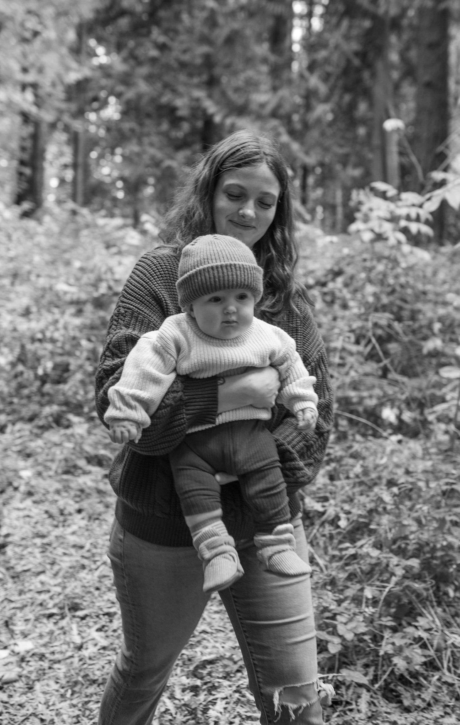 Black and white photo of a young woman holding a baby in a knit hat outdoors in a lush forest setting.