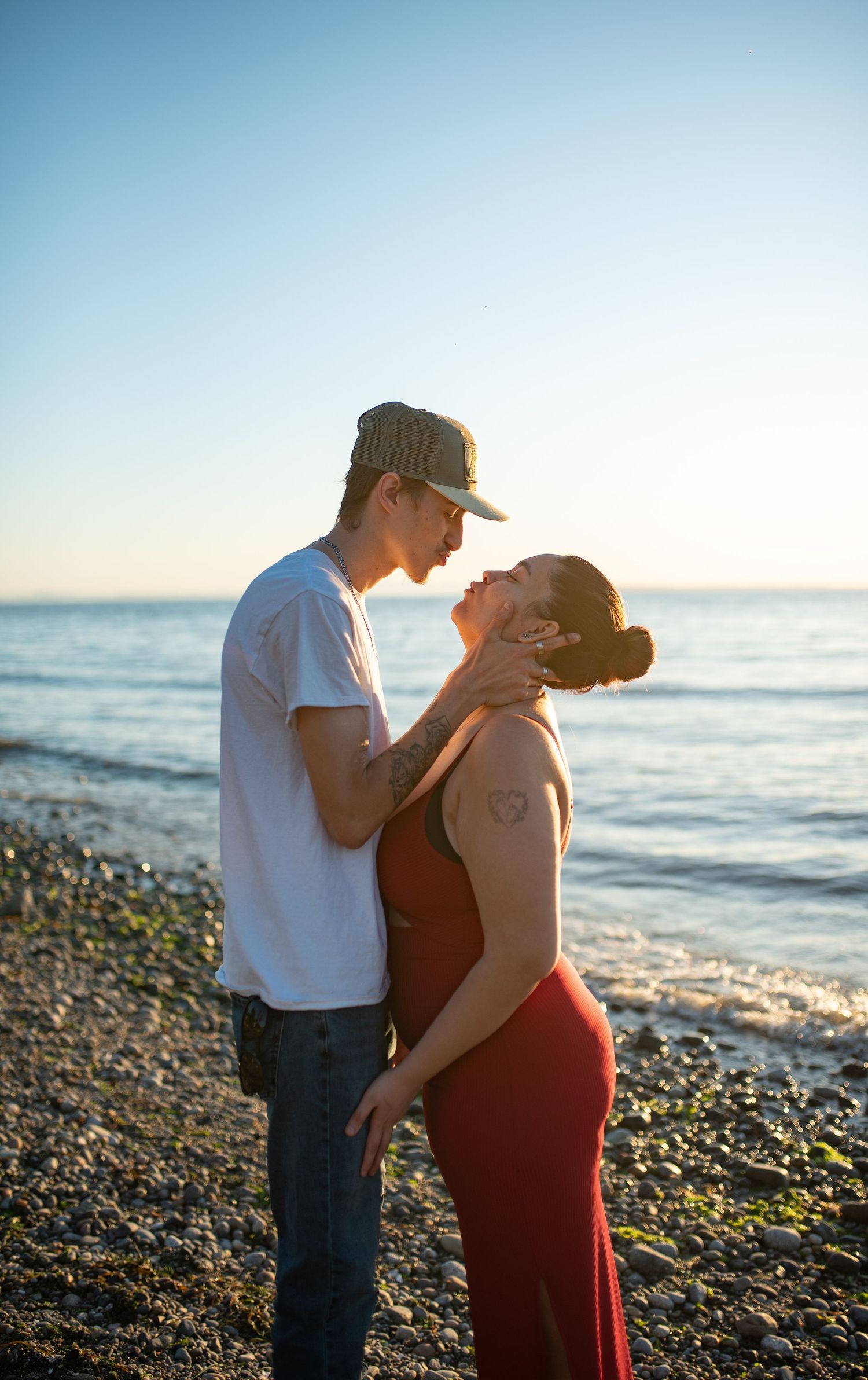 Romantic couple almost kissing on rocky beach at golden hour, man in cap holding woman's face.