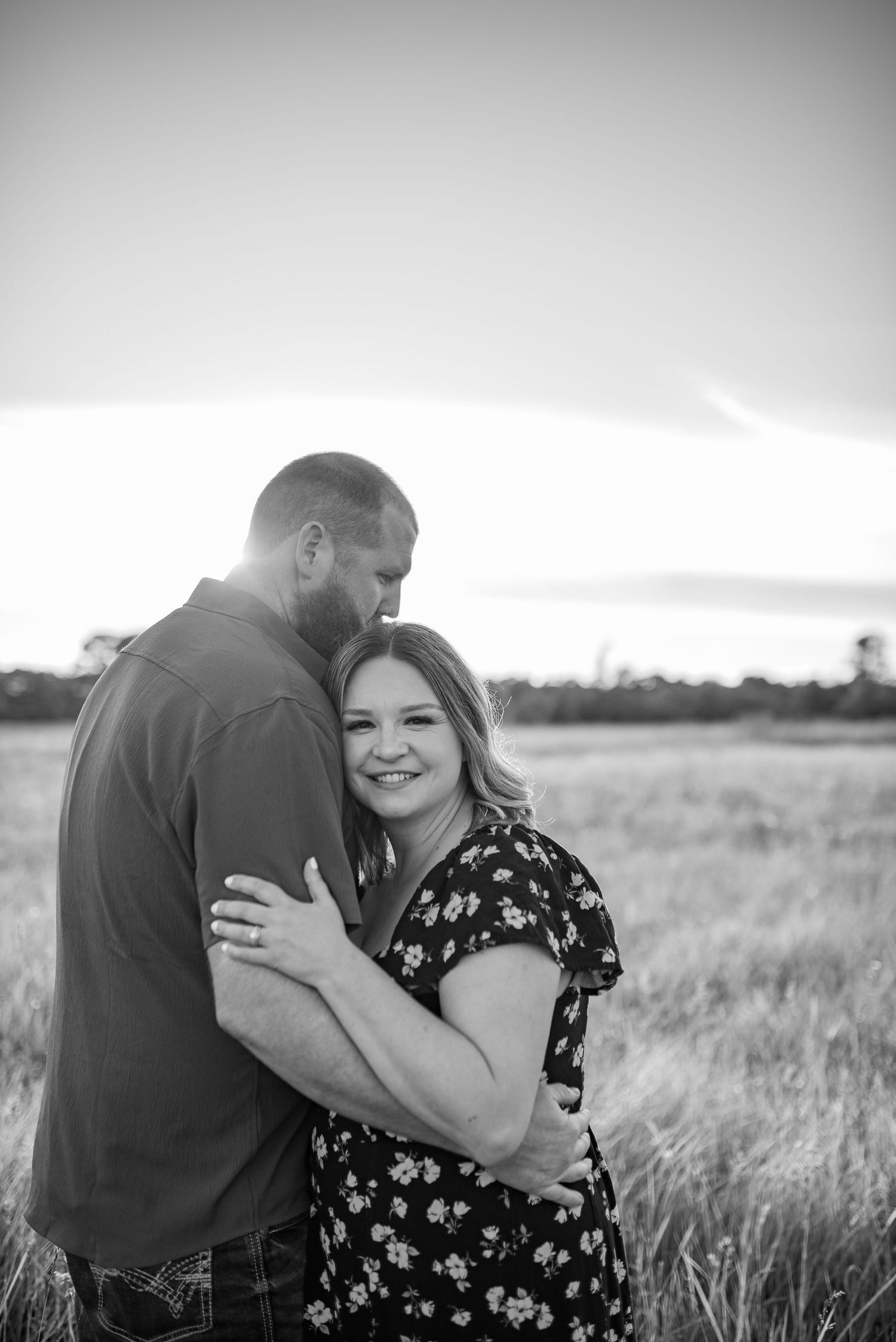 Black and white photo of a smiling couple embracing in an open field at sunset, woman in floral dress.