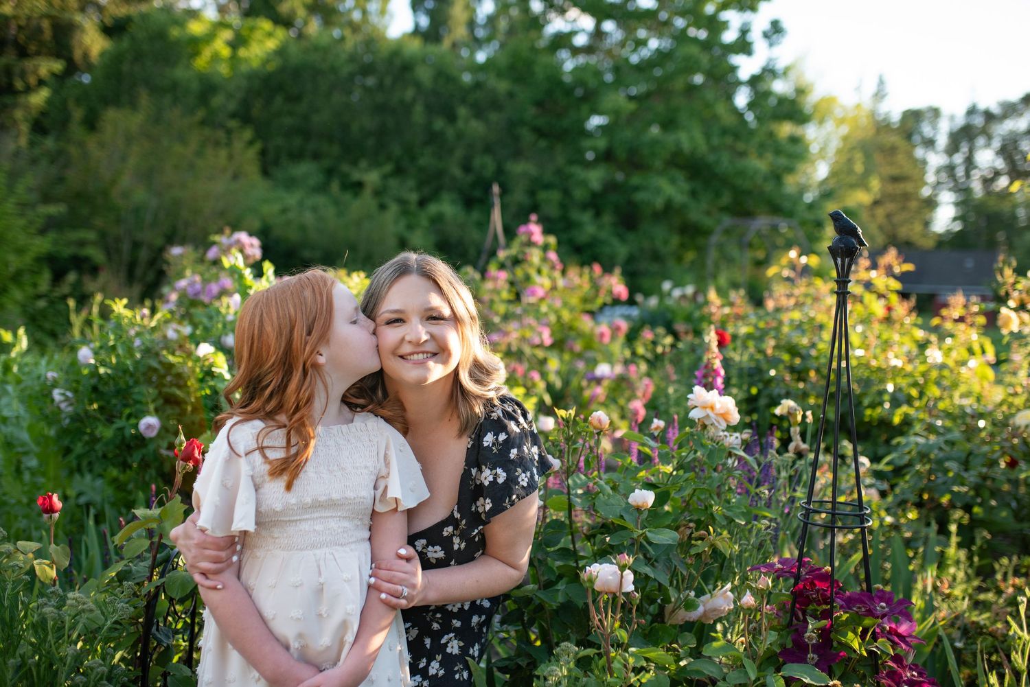 Young red-haired girl kisses smiling blonde woman on cheek in a lush flower garden at golden hour.