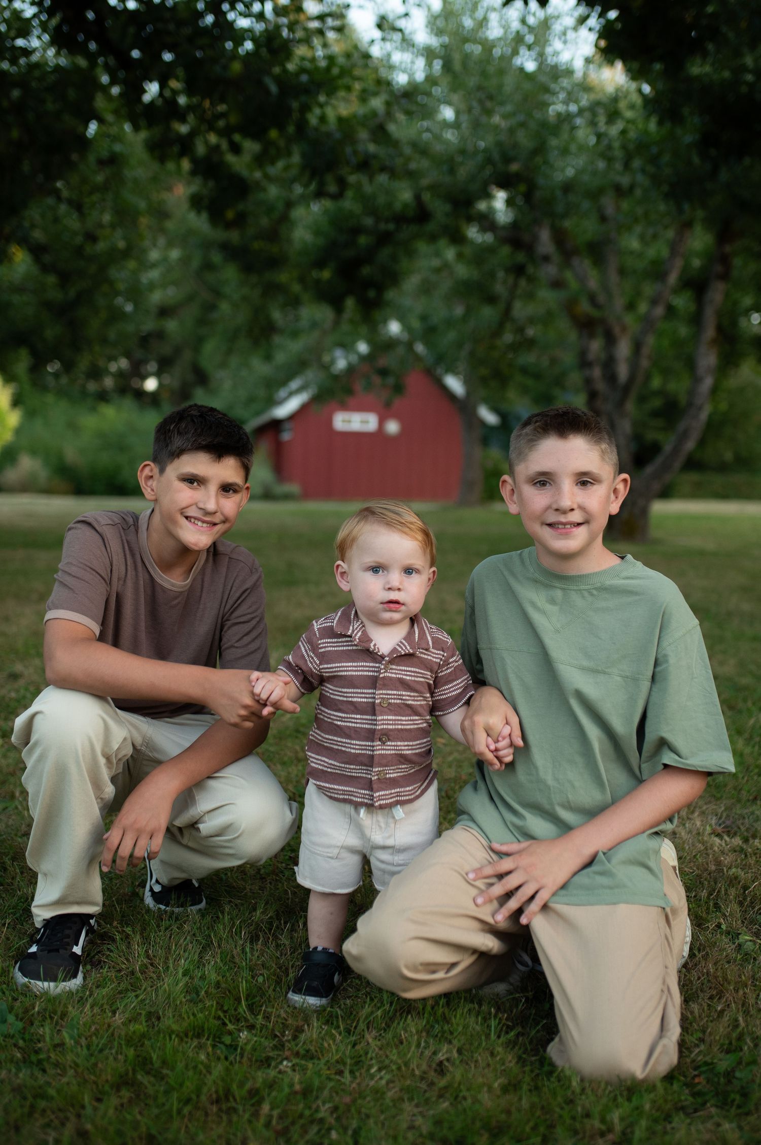 Three brothers posing outdoors with a red barn background, two older boys kneeling beside a smiling toddler.