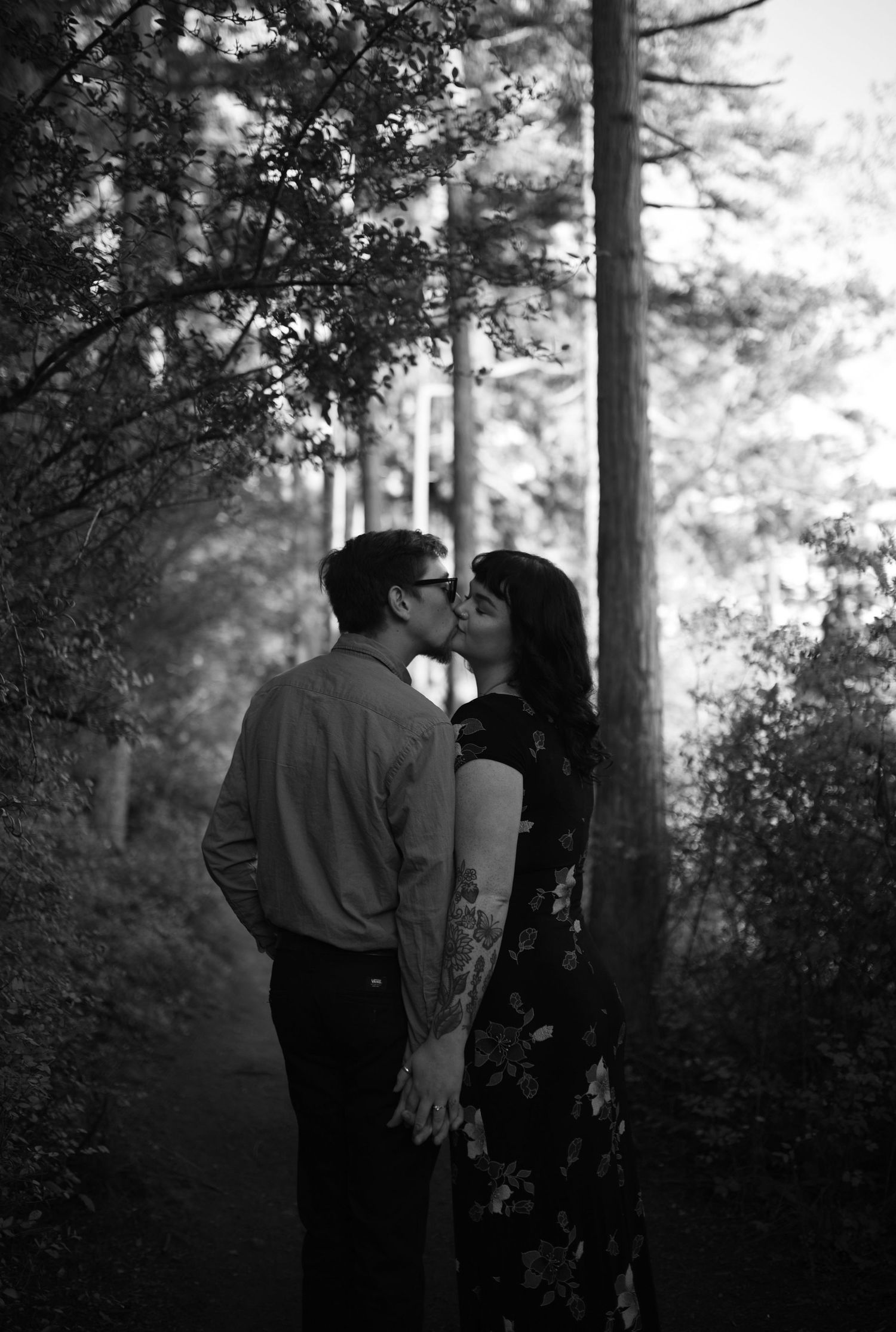 Black and white photo of couple sharing a kiss in a misty forest pathway.