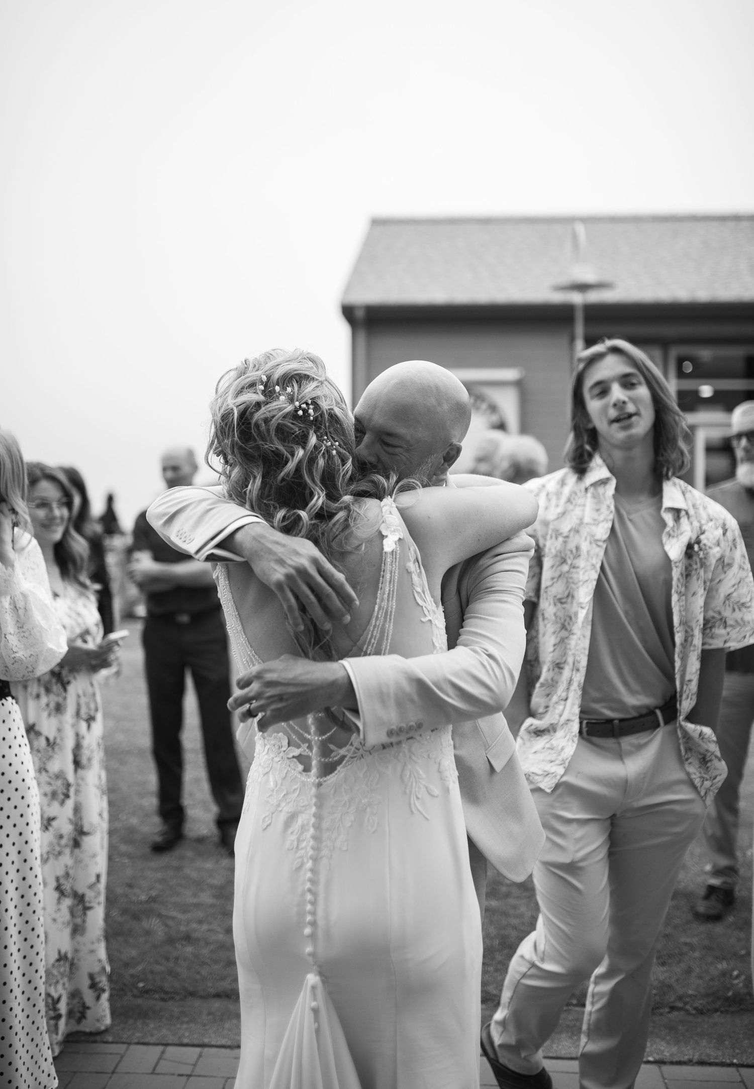 Black and white wedding photo of bride and groom embracing outdoors, guests watching nearby.