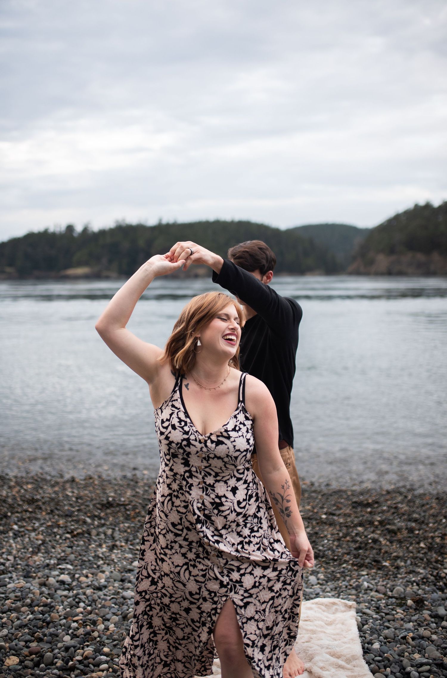 Woman in floral dress laughing as her partner spins her on a rocky Pacific Northwest beach at dusk.