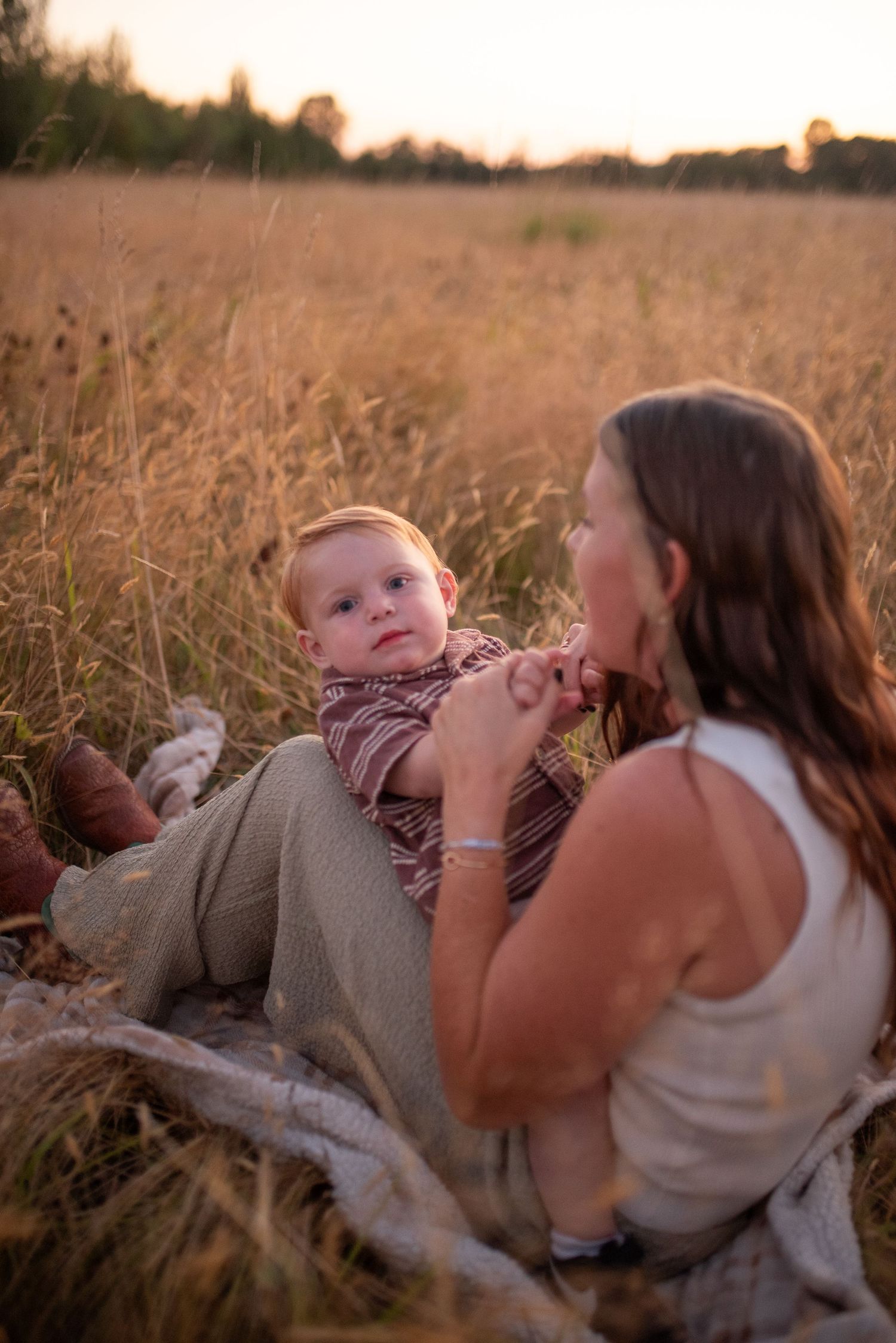 Mother sitting in golden field at sunset, tenderly interacting with her baby on a blanket.