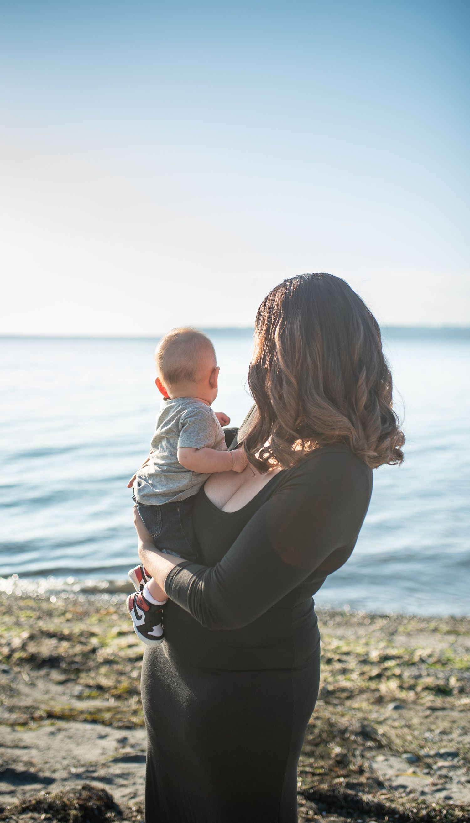 Mother standing on rocky shoreline holding her baby while gazing out at the calm ocean.