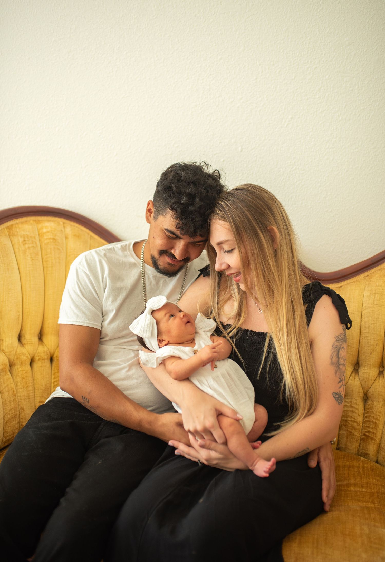 Young couple leaning heads together lovingly while holding their newborn baby on a gold sofa.