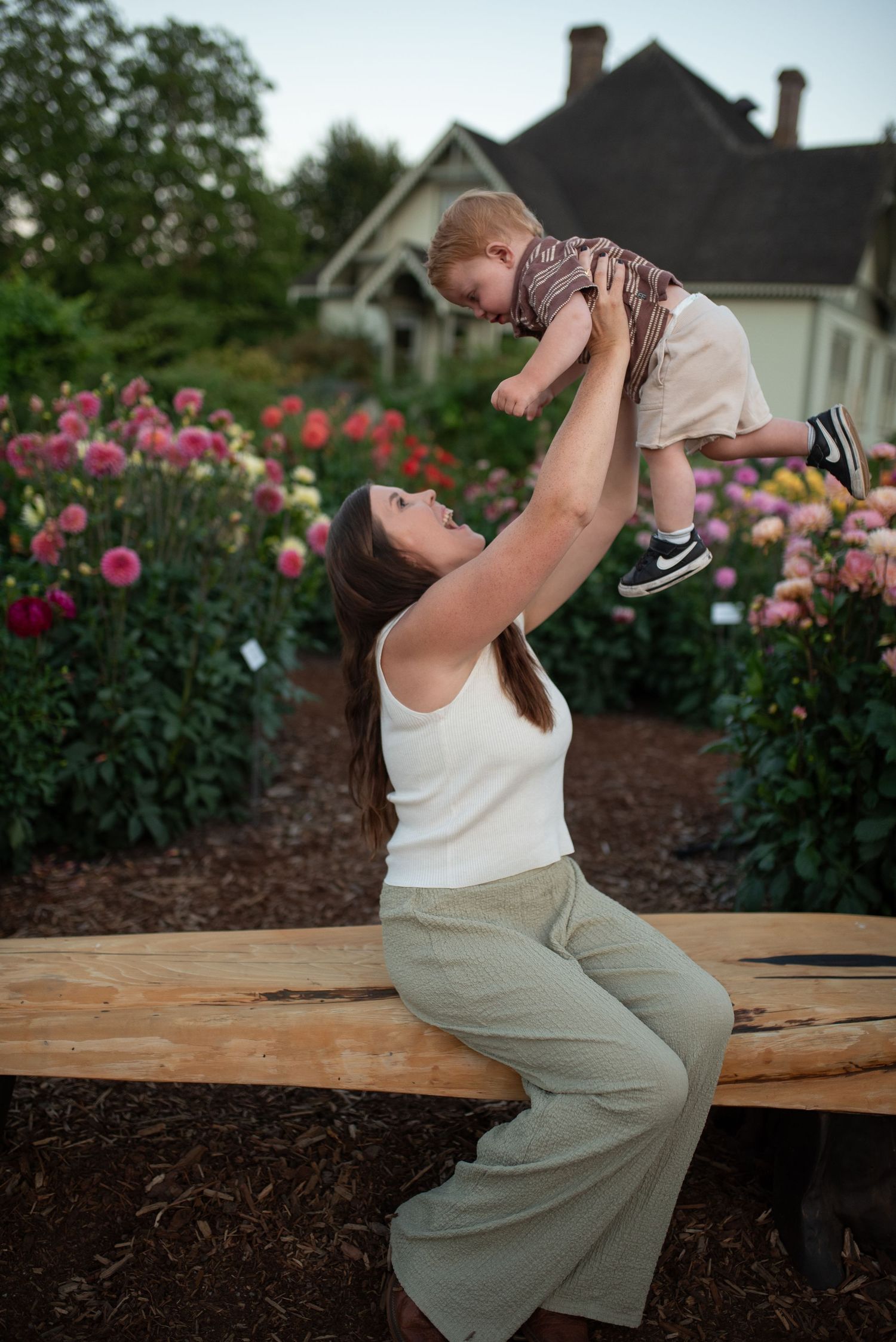 A woman sits on a wooden bench in a flower garden, joyfully lifting a toddler above her head.