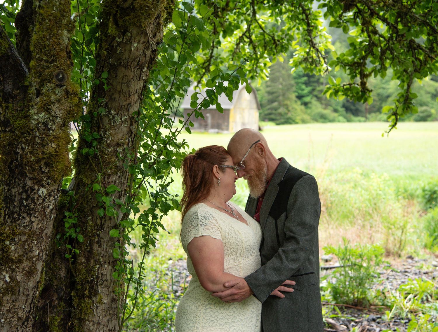 Couple sharing a romantic moment under a leafy tree in a lush green field, dressed in wedding attire.