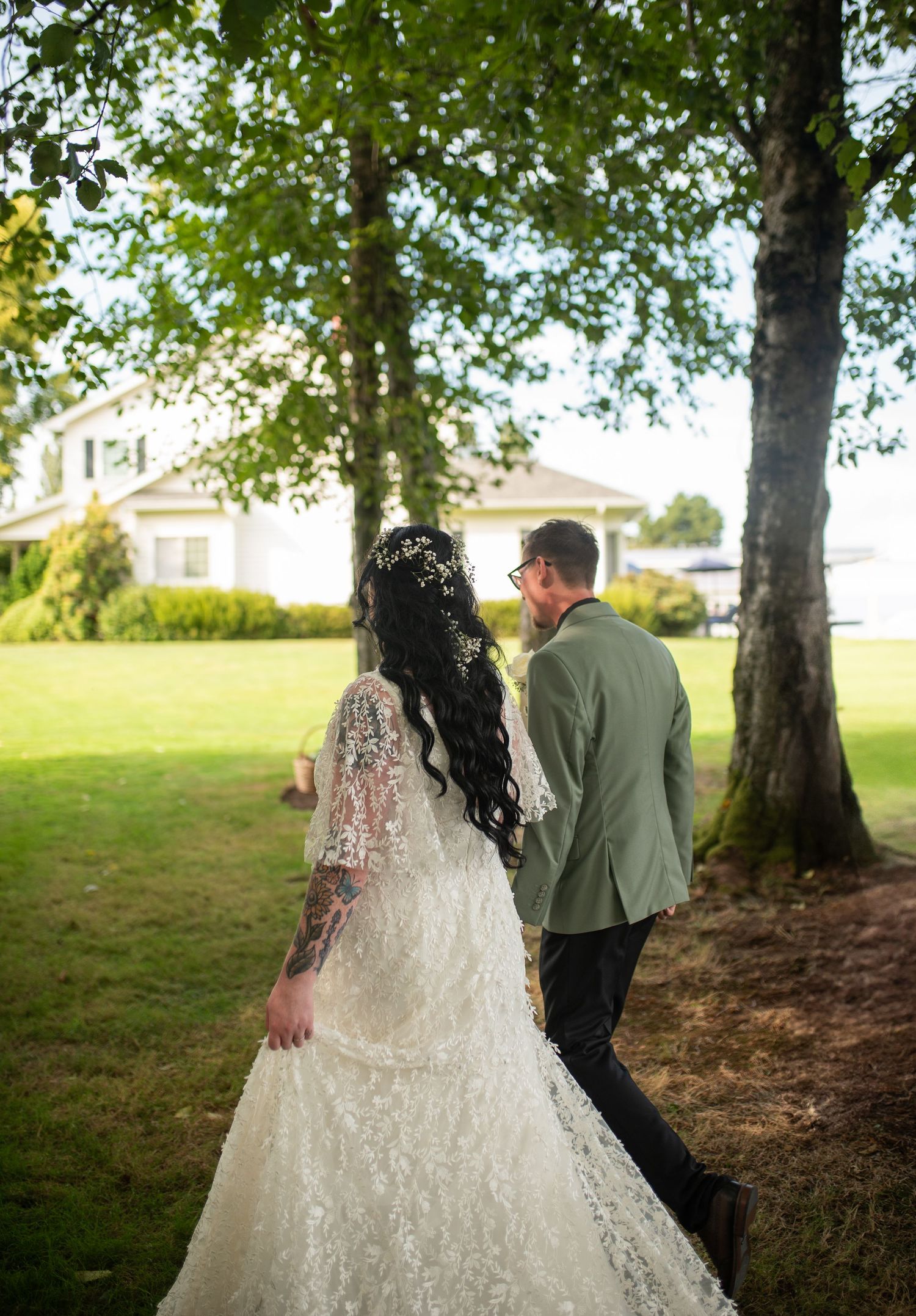 Bride in lace gown and groom in green suit walking hand in hand under trees at outdoor wedding.
