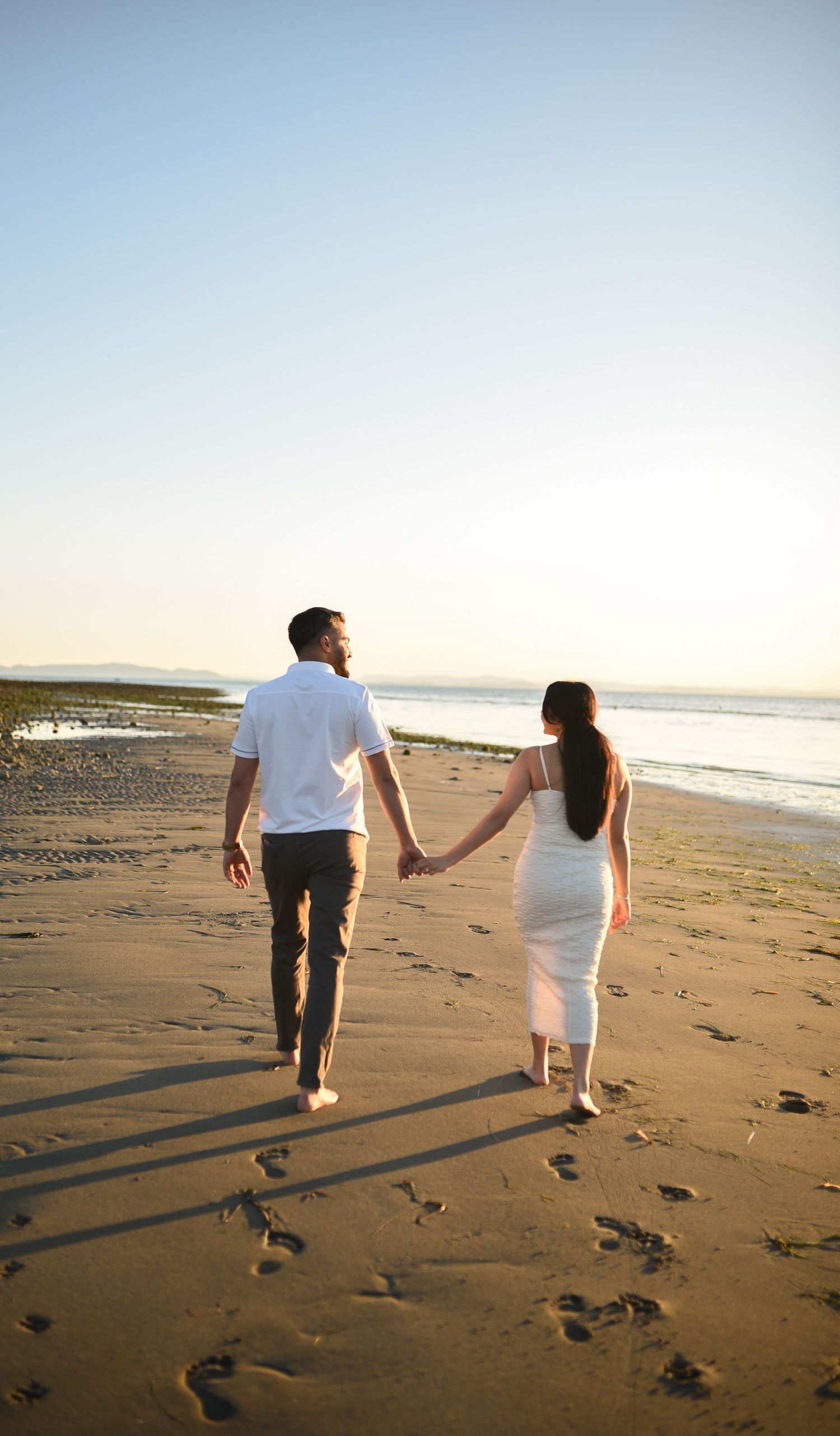 Couple holding hands walking barefoot on a sandy beach at sunset, woman in white dress and man in casual attire.