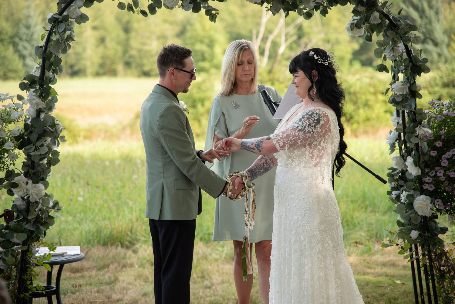 Couple exchanging vows at outdoor wedding ceremony under floral arch with officiant in green field.