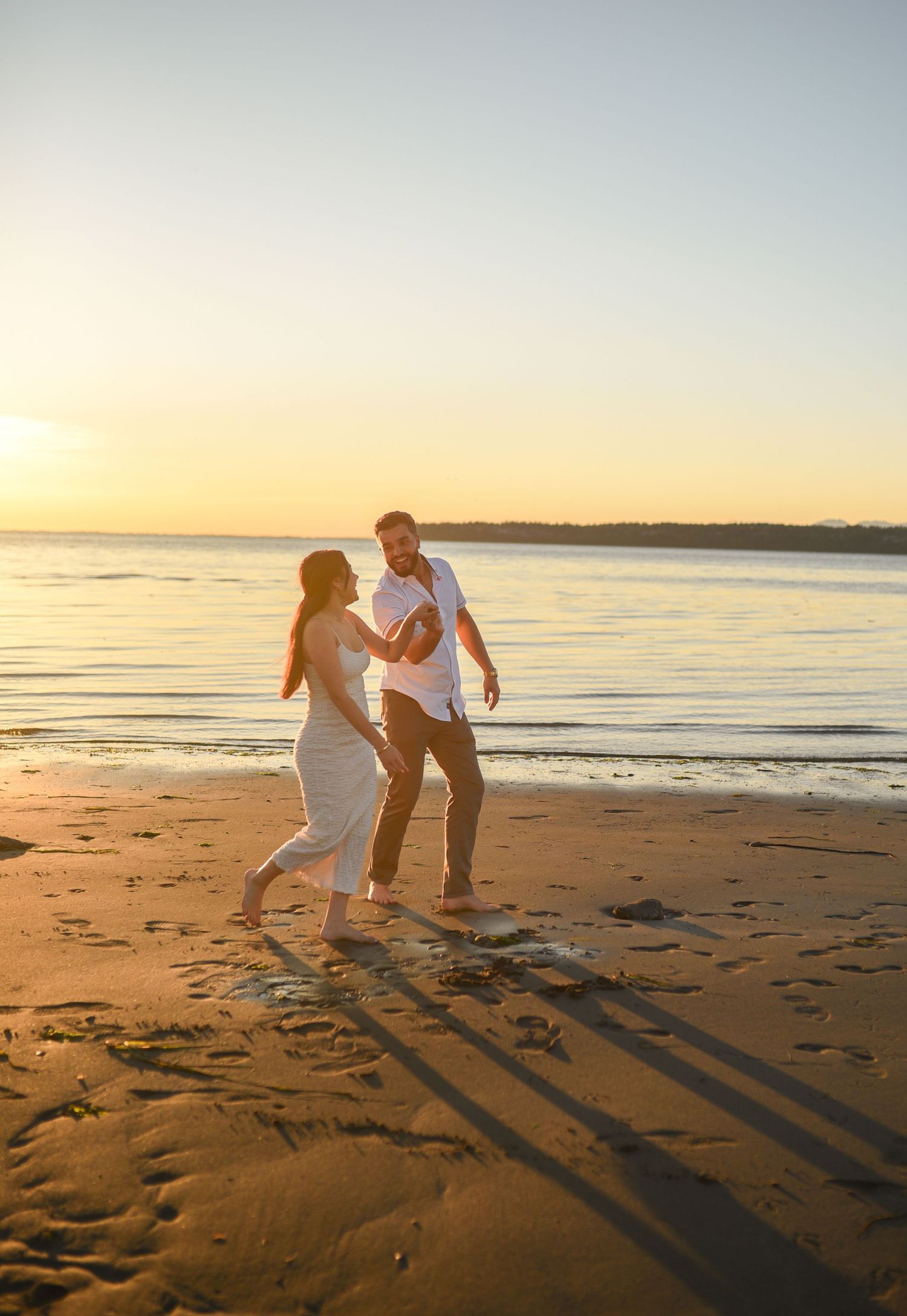 Couple dancing joyfully on a sandy beach at golden sunset, waves reflecting warm light behind them.