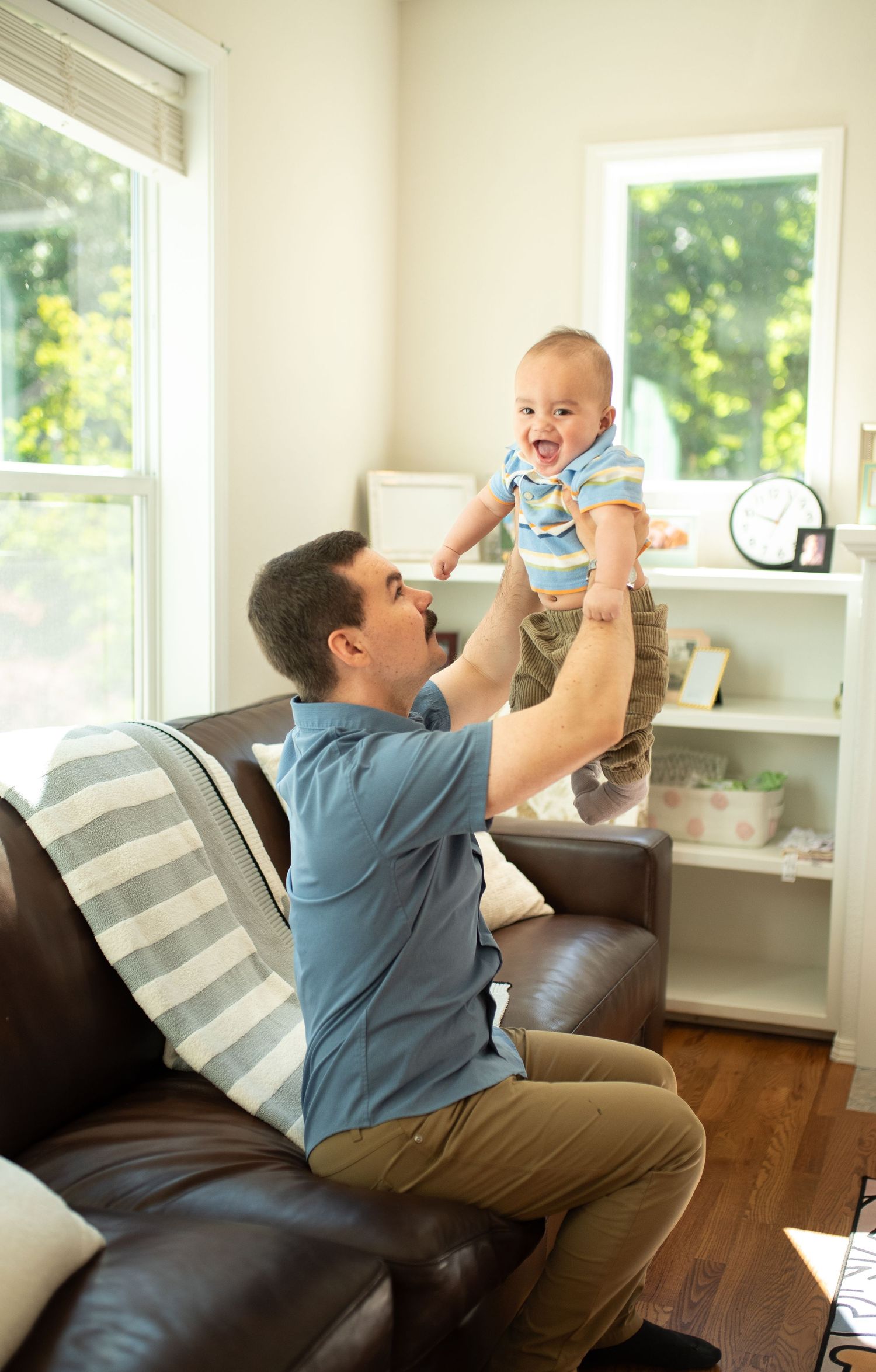 Father joyfully lifting laughing baby in sunny living room.