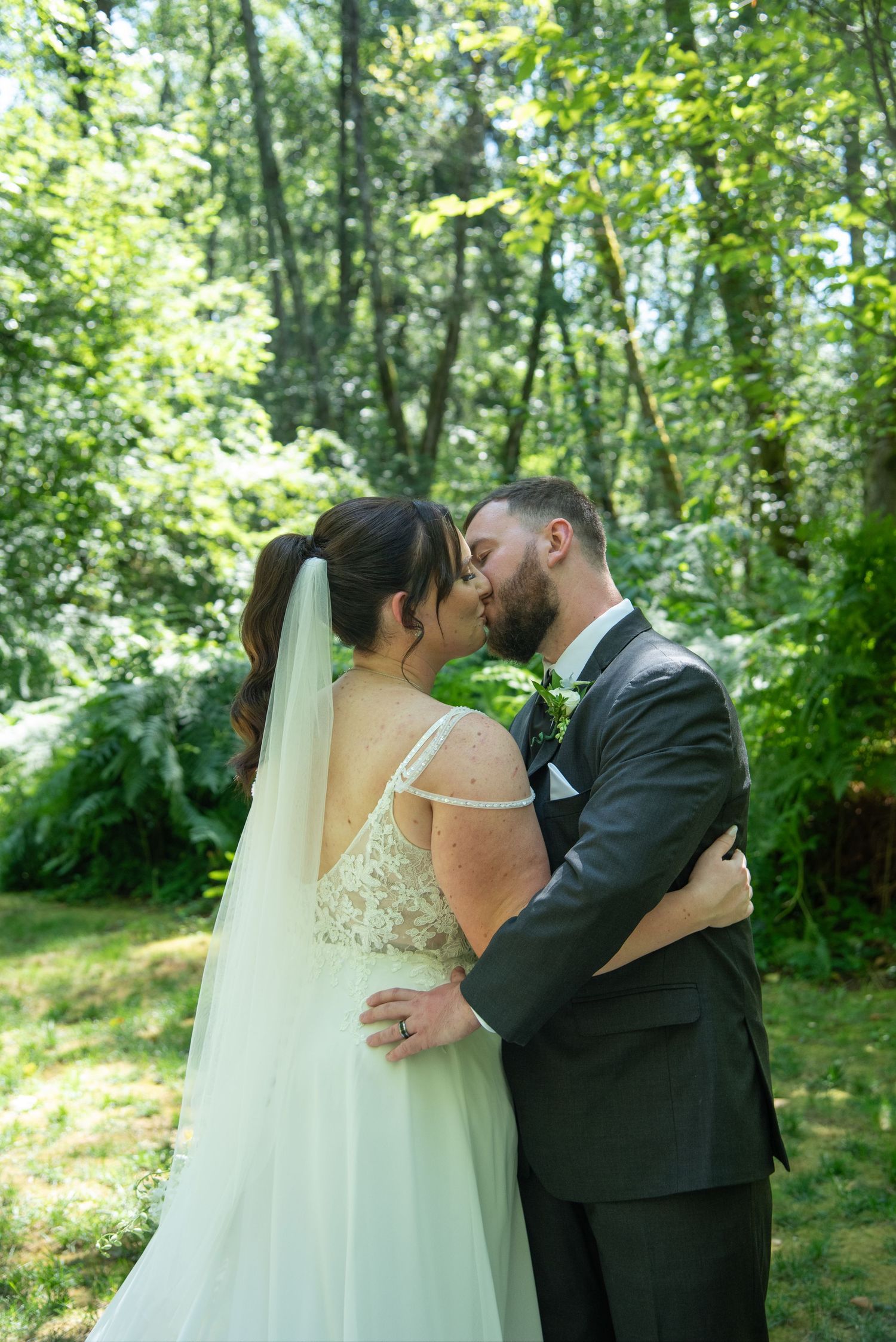 A bride and groom share a romantic kiss surrounded by lush green trees in a forest setting.