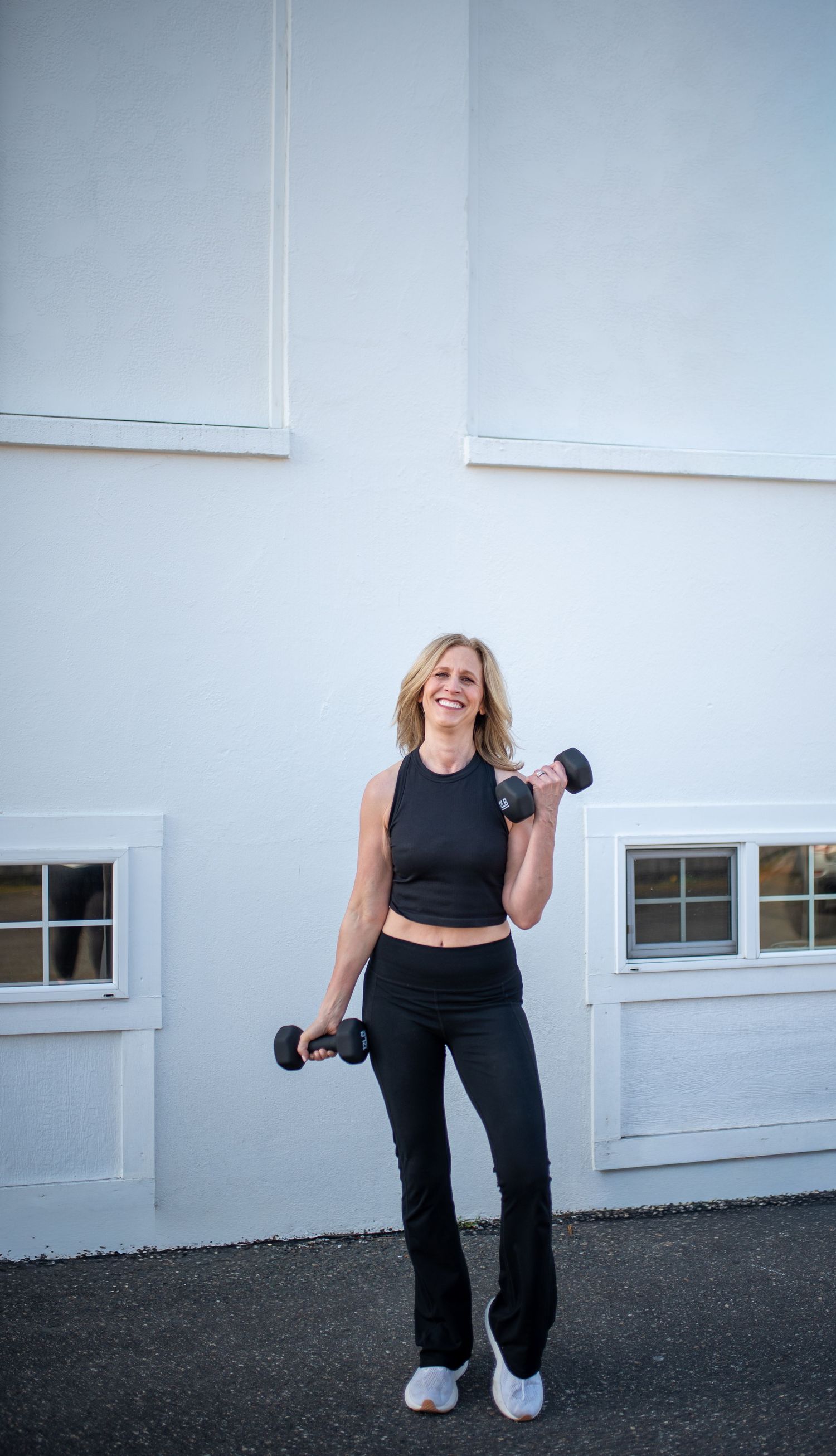 Smiling blonde woman in black workout outfit holds dumbbells outdoors against a white building wall.