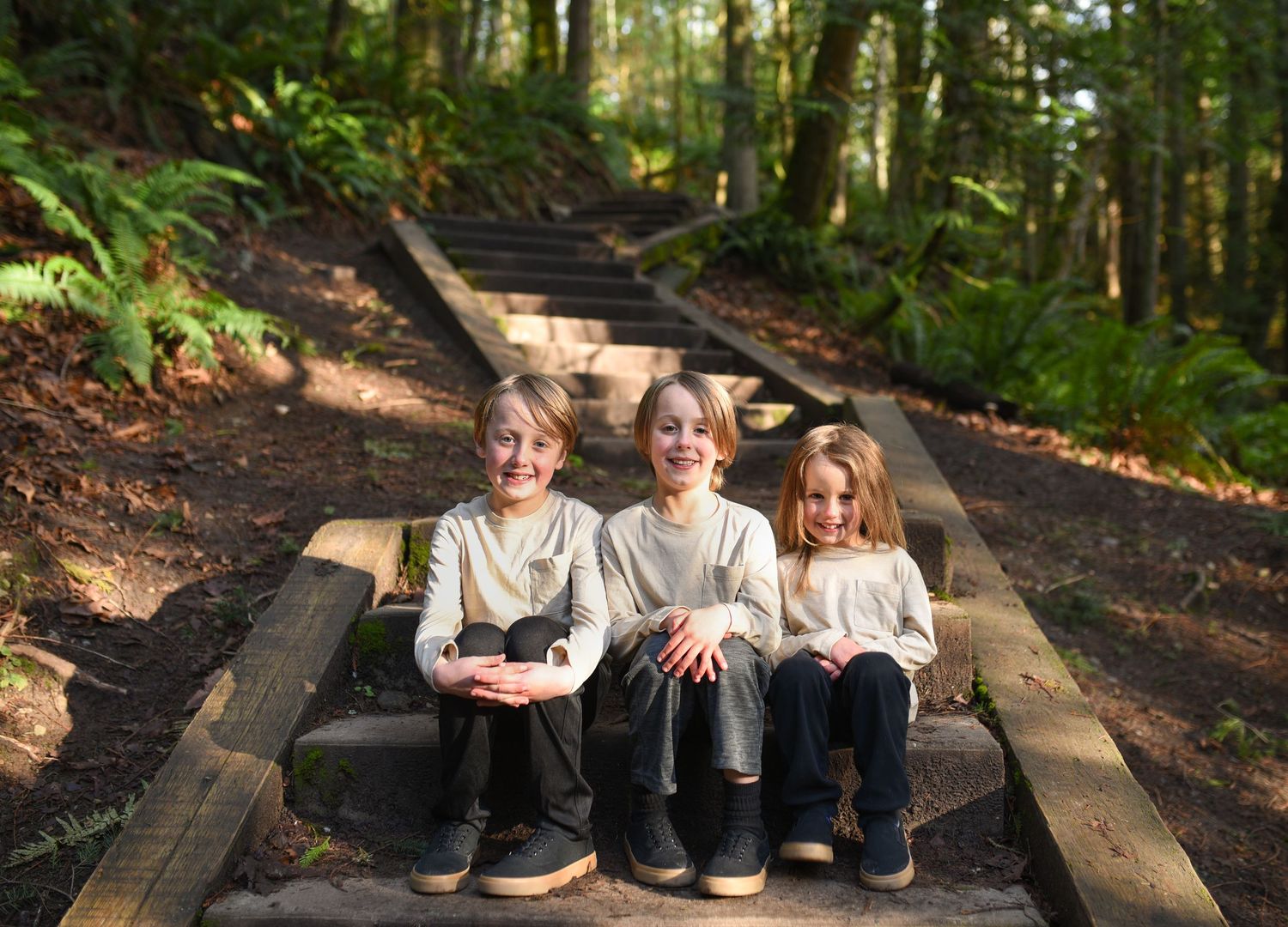 Three smiling children sit together on wooden steps surrounded by lush green forest foliage.
