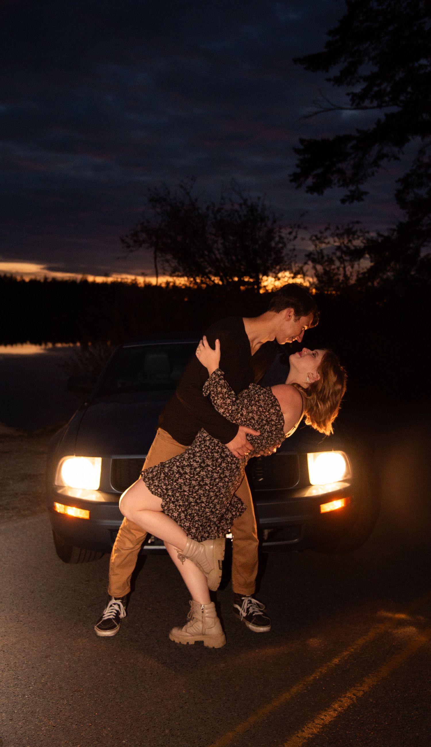Couple sharing a dramatic dip kiss in front of a classic car with glowing headlights at twilight.
