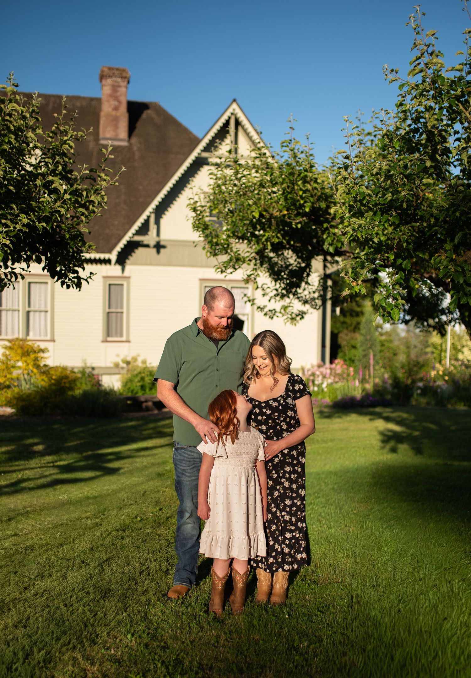 Family of three sharing tender moment on green lawn before historic home.